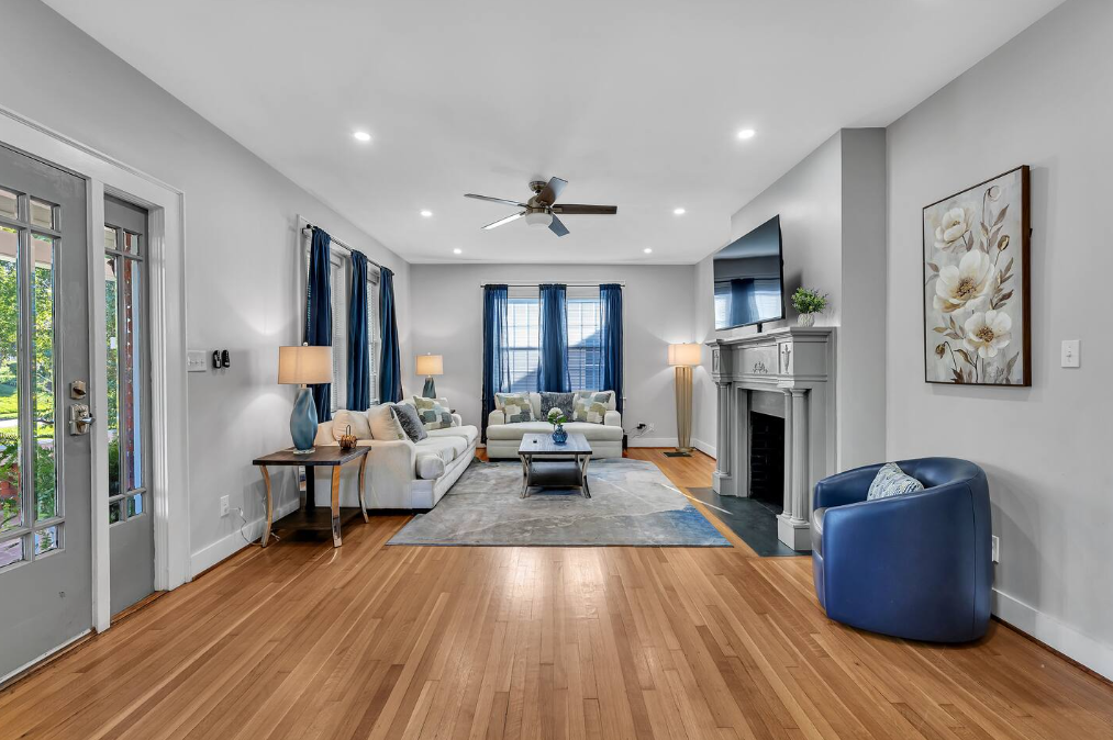 Bright living room with white walls, hardwood floors, a gray fireplace with a mounted TV, blue curtains, and multiple sofas with decorative pillows. There are two lamps, a ceiling fan, and artwork of white flowers on the wall.