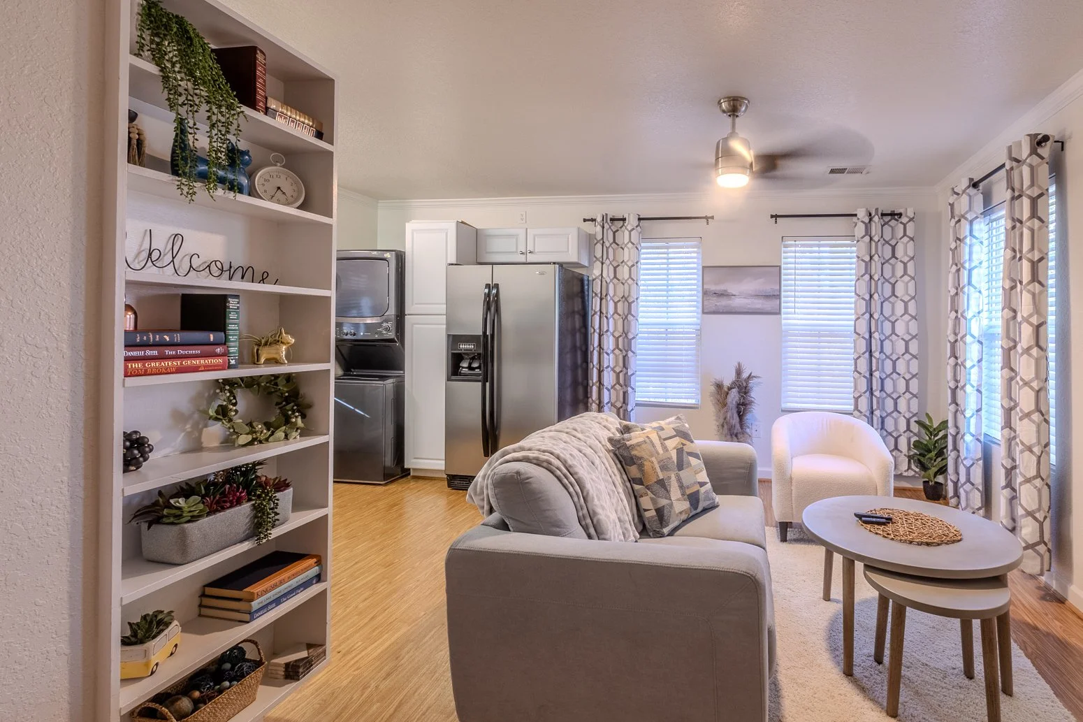 Living room with white walls and hardwood floors, featuring a light gray sofa and chairs, a white oval coffee table, decorative pillows, a bookshelf with various decorations, and a kitchen area with a stainless steel refrigerator and white cabinets i