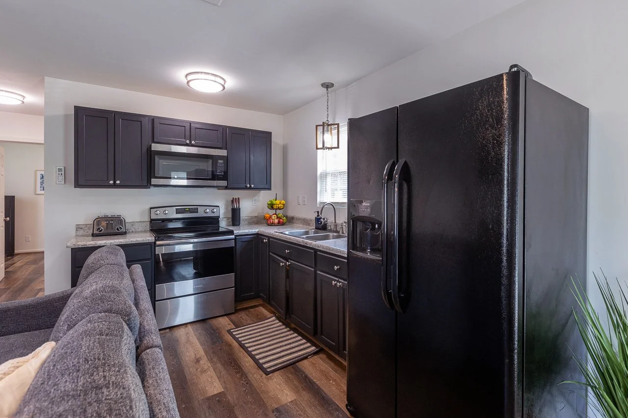 Kitchen with dark cabinets, black refrigerator, stainless steel appliances, and a gray couch partially visible in the foreground.