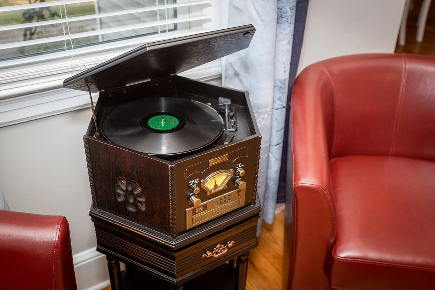 Vintage wooden record player with open lid, placed beside a red leather armchair near a window with blinds.
