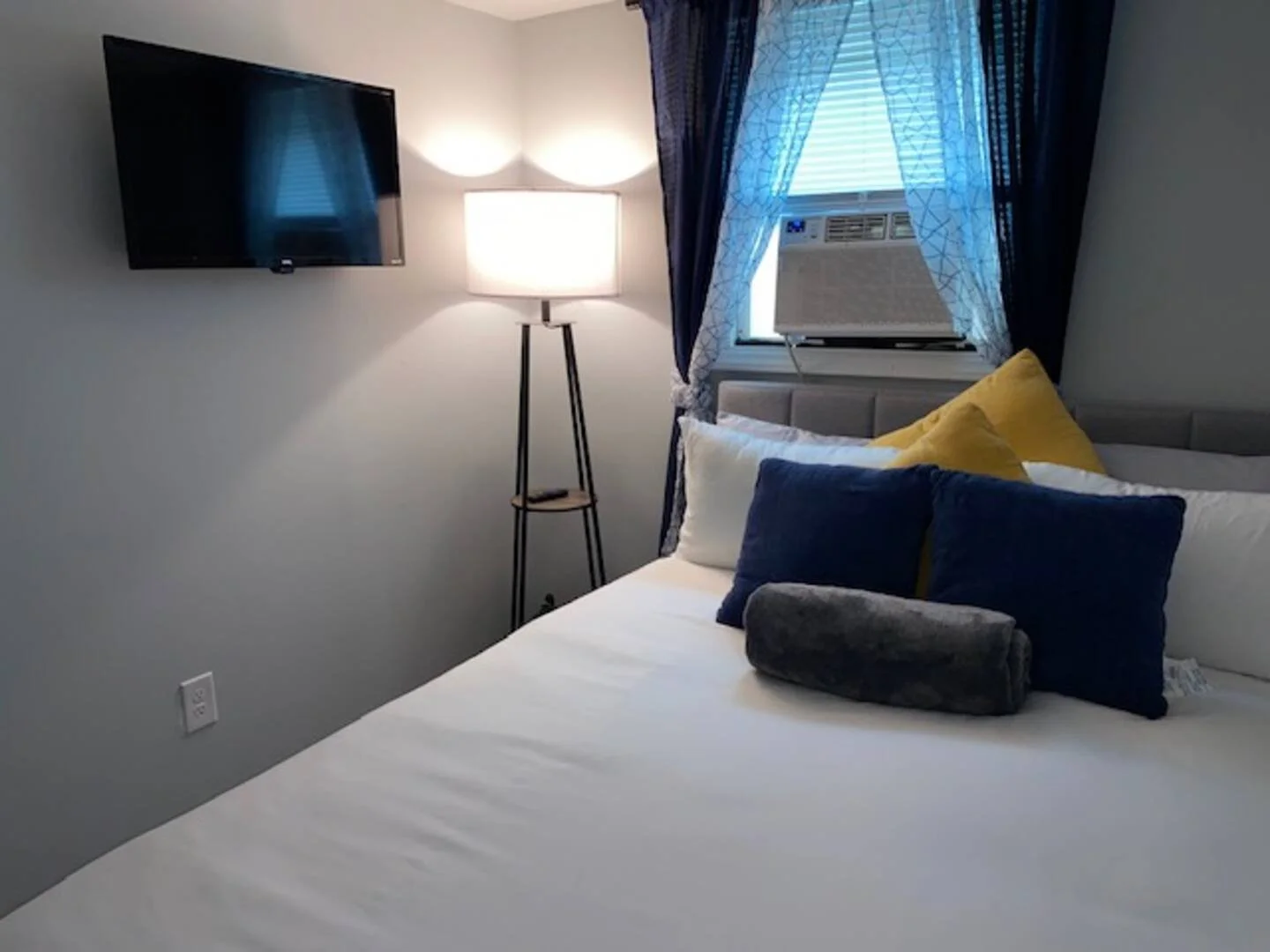 A bedroom with a white bed, yellow and blue pillows, a window with blue curtains, an air conditioner, a wall-mounted TV, and a standing lamp.