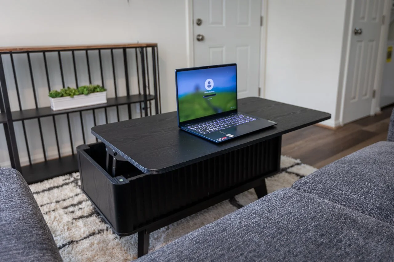 Living room with a black coffee table, gray sofas, a black bookshelf with a plant, and a closed white door in the background. A laptop is on the coffee table.