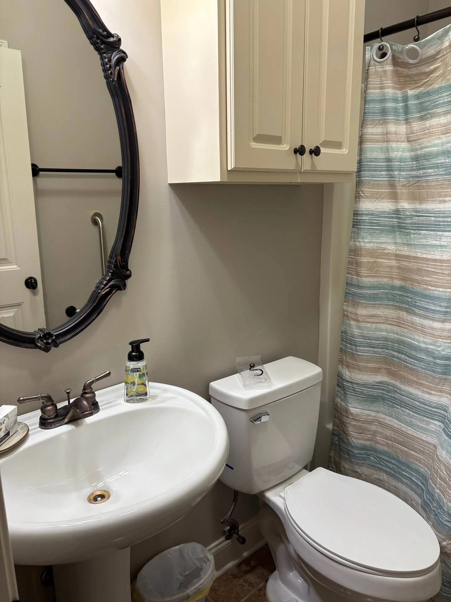 A small bathroom with a white sink, a mirror with a dark ornate frame, a toilet with a plastic shield on top, a beige cabinet above, a striped shower curtain, and a small trash can underneath the sink.