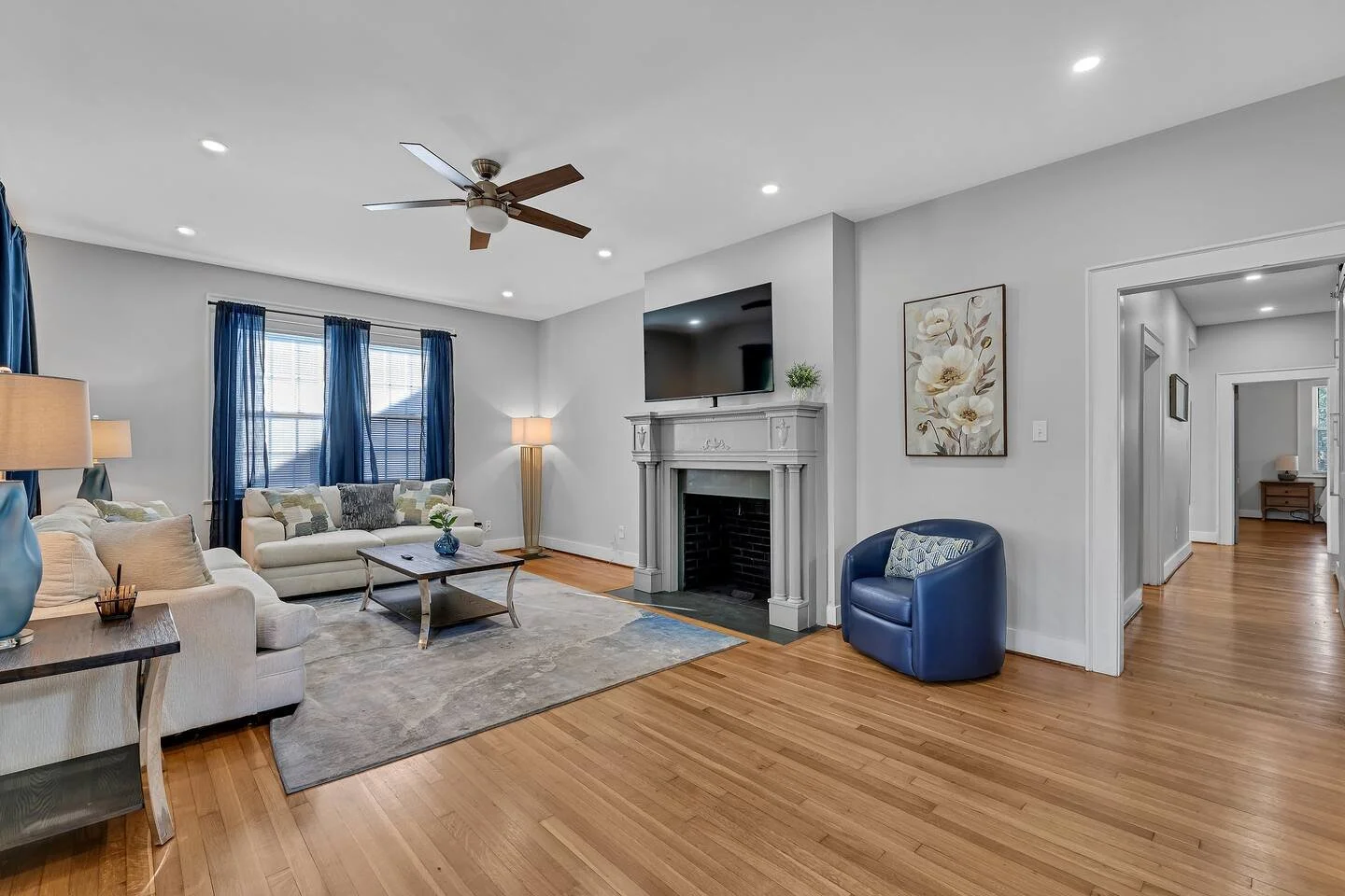 Living room with light-colored sofas, dark blue curtains, blue accent chair, fireplace with a TV on top, hardwood floors, a rug, and artwork on the wall.