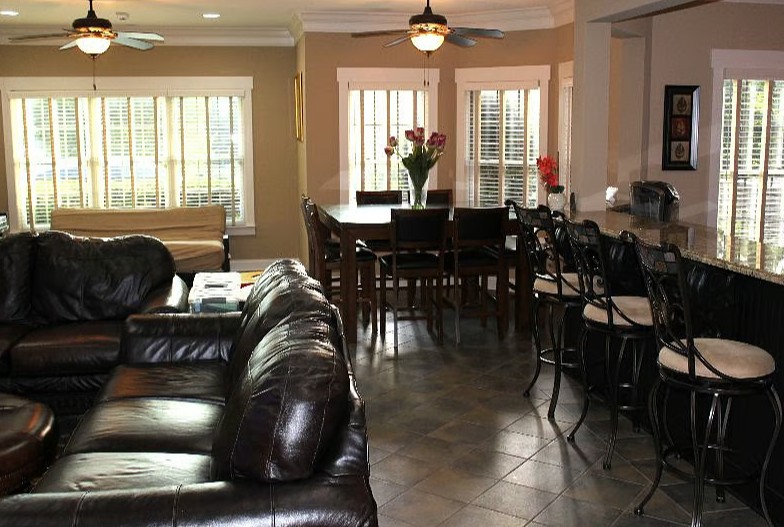 Living and dining room with leather sofa, wooden dining table with flowers, bar area with high chairs, large windows with blinds, ceiling fans, tiled floor.