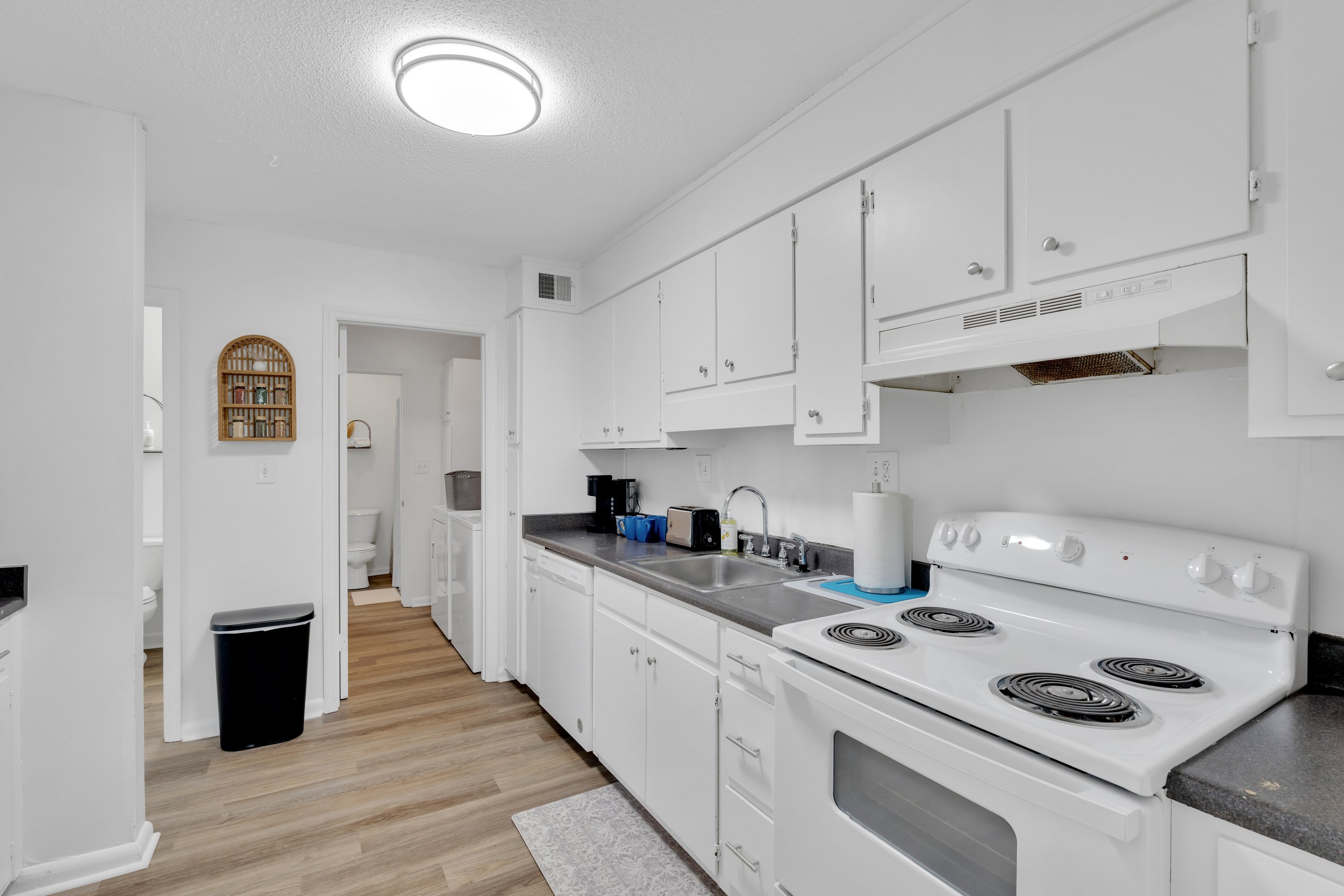 Kitchen with white cabinets, black countertop, white stove, stainless steel sink, paper towel roll, coffee machine, and small appliances on the counter. A doorway shows a laundry area with washer and dryer and a bathroom with a toilet.
