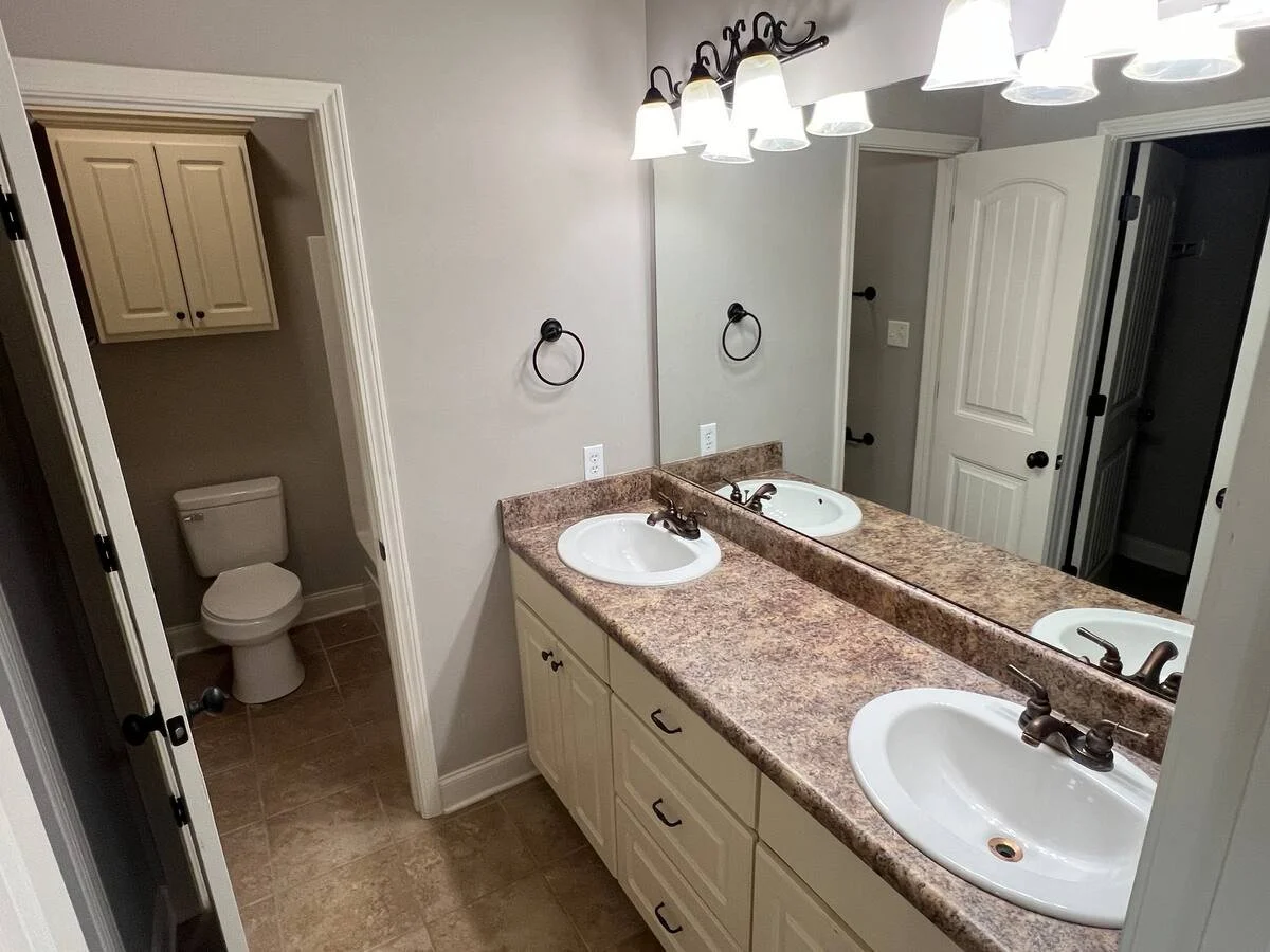 Bathroom with a double sink vanity with a large mirror, a light fixture above, and a separate toilet area visible through an open door.
