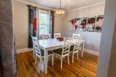 A dining room with a white table and six matching chairs, a large window with curtains, a decorative wall art, and hardwood flooring.