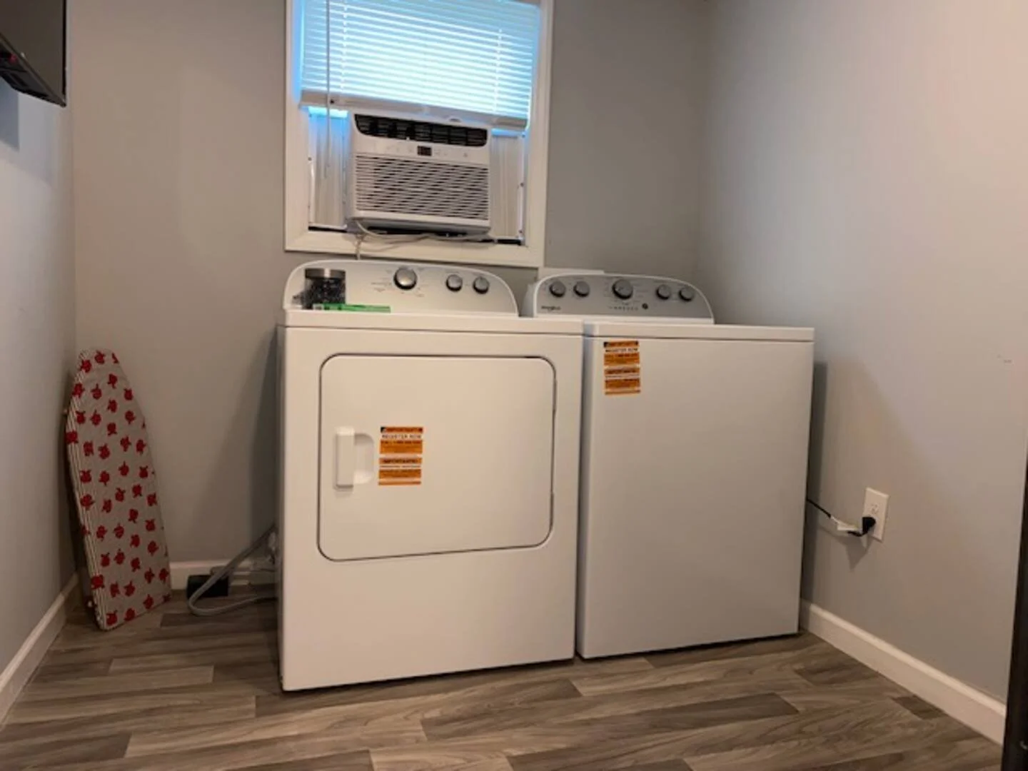 A laundry area with a white top-loading washing machine and a white dryer side by side against a wall. There is a small window above them with an air conditioning unit installed. An iron rests against the wall near the appliances, and a power outlet 