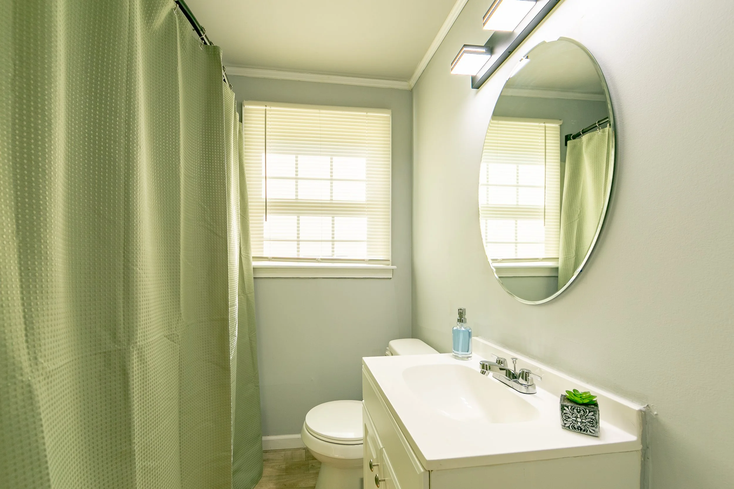 A small bathroom with a light green shower curtain, a white vanity with a sink, a round mirror above the sink, a window with blinds, a bottle of soap, and a decorative plant on the vanity.