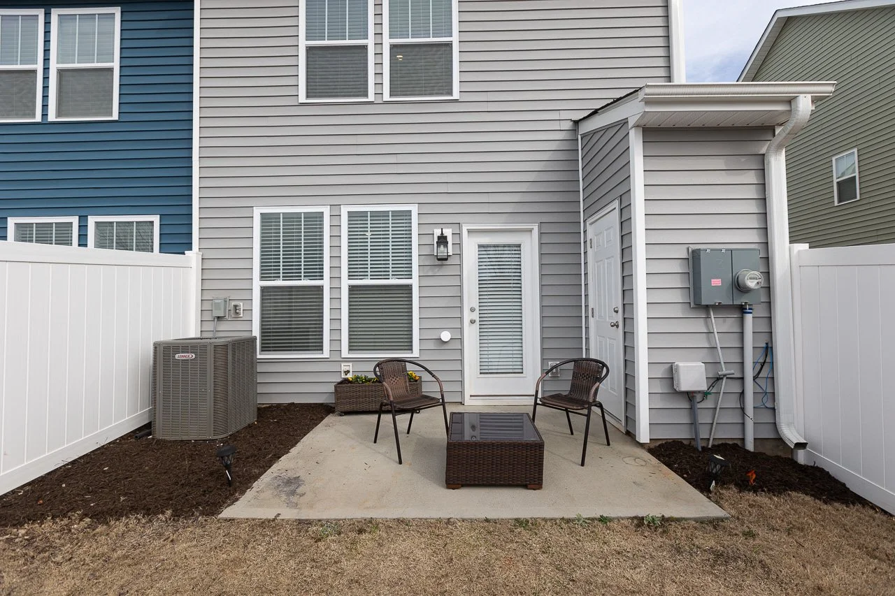 Backyard patio area with a concrete slab, two metal chairs, a wicker coffee table, a flower box, a gray house wall with windows, a door with blinds, outdoor lighting, and HVAC equipment enclosed by white fences.