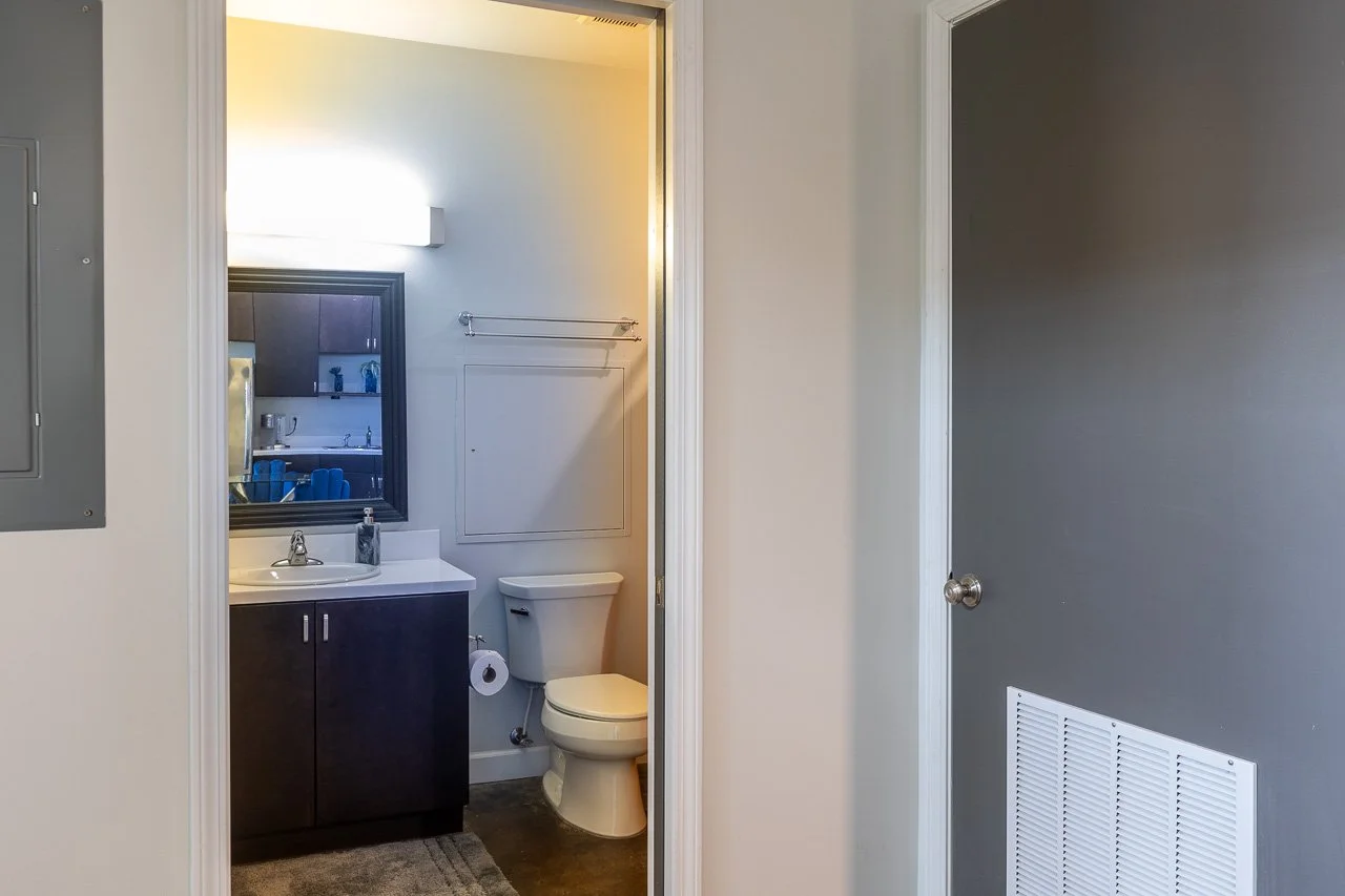 A small bathroom with a dark vanity, a mirror, a sink, a toilet, and a towel rack, seen through a doorway from a room with a gray wall and a ventilation vent.