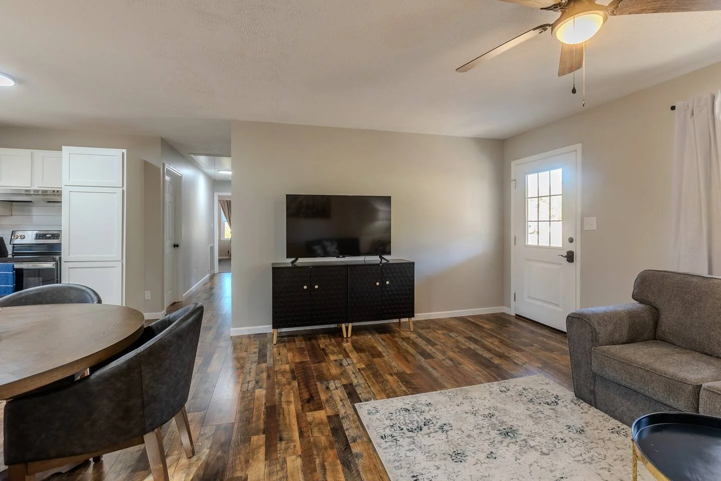 Living room with hardwood floors, a gray sofa, a patterned area rug, a black TV stand with a TV, a front door with a glass window, and a ceiling fan with a light, with a view into the kitchen and hallway.
