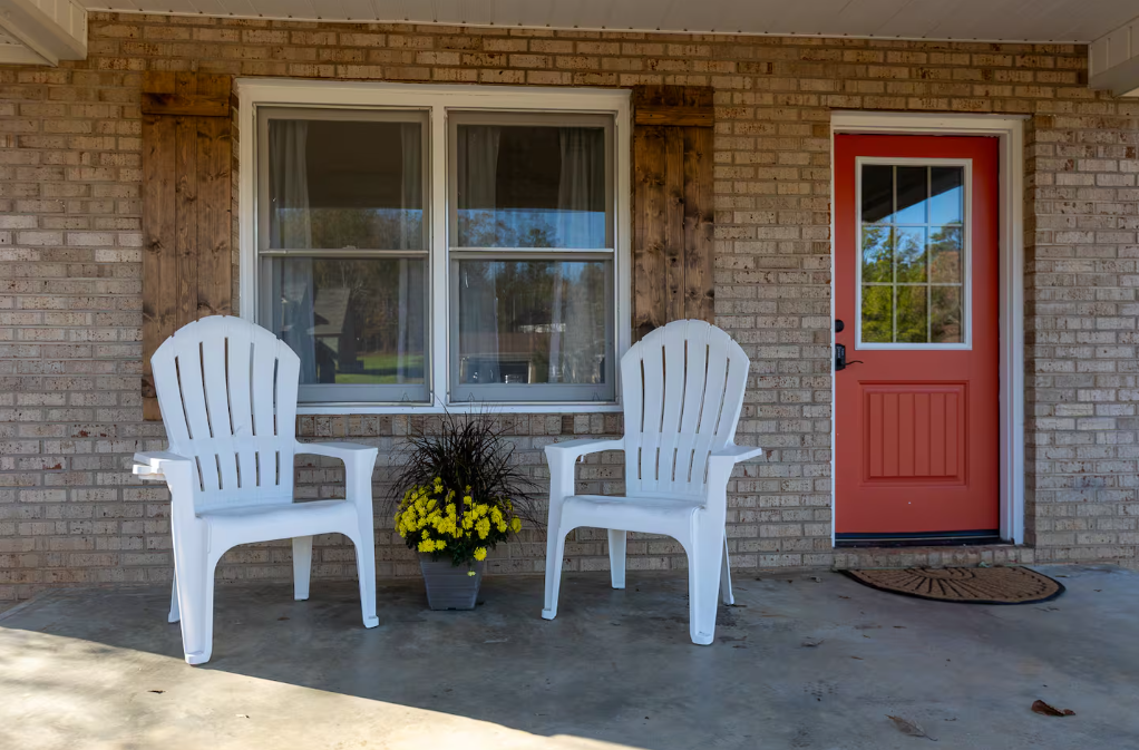 Two white Adirondack chairs and a potted yellow chrysanthemum flower on the porch outside a brick house with a red door and window.
