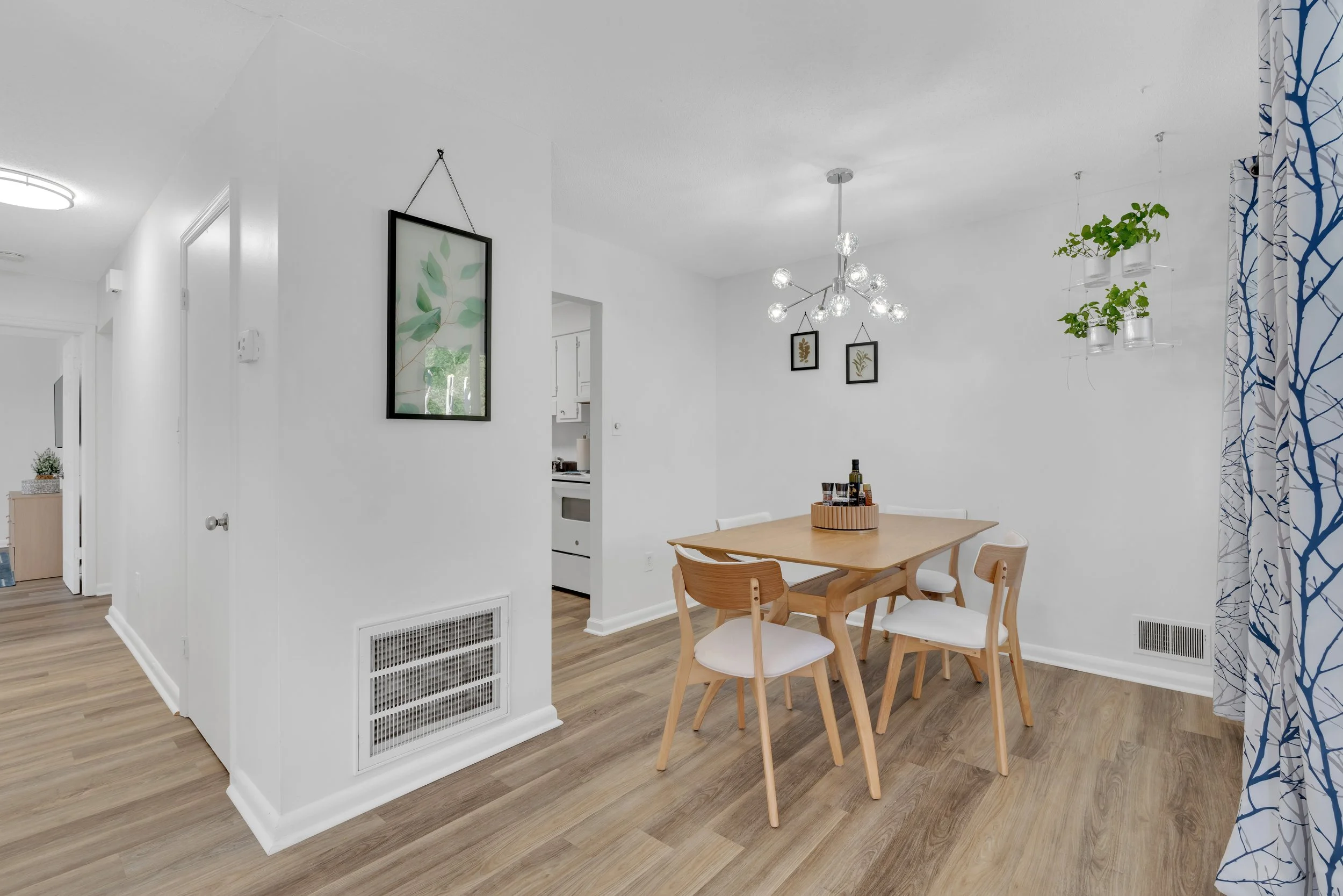 Dining area with a wooden table and four chairs, modern chandelier, white walls with framed botanical art, and hanging plants near a window with patterned curtains.