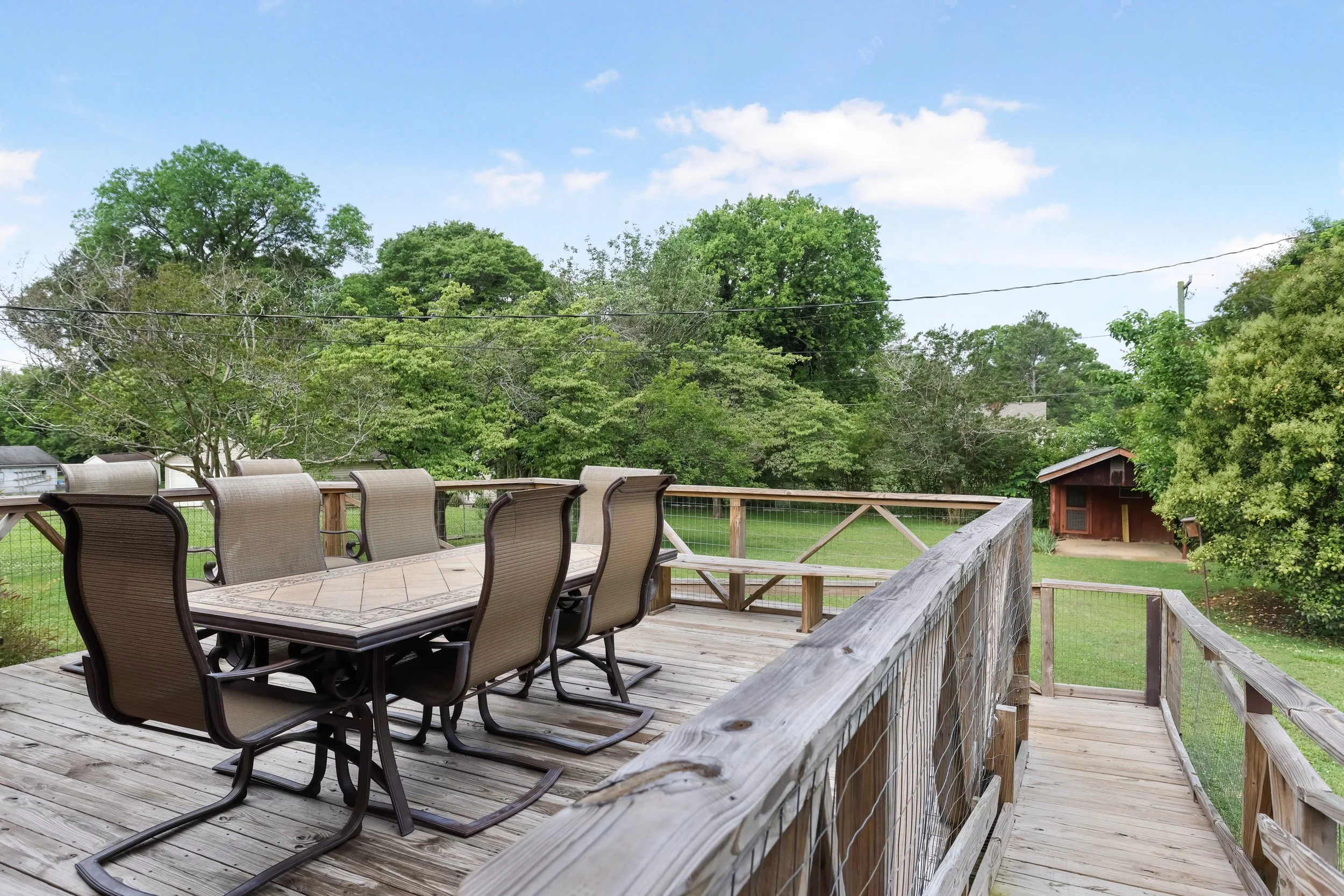 Wooden outdoor deck with patio table and six chairs, overlooking a lush green backyard with trees and an outbuilding.