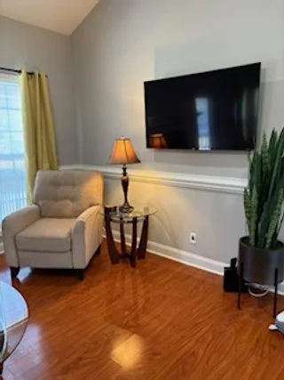 Living room corner with a beige armchair, a wooden side table with a lamp, a large wall-mounted TV, a tall green potted plant, and yellow curtains on a window.