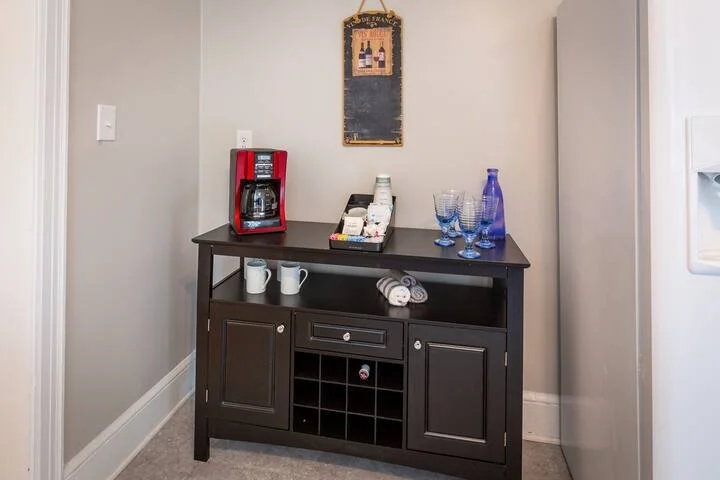 A black wooden sideboard with a drawer, closed cabinets, and open wine cubbies, holding a coffee maker, mugs, napkins, and glassware, located against a beige wall with a chalkboard and electrical outlets.