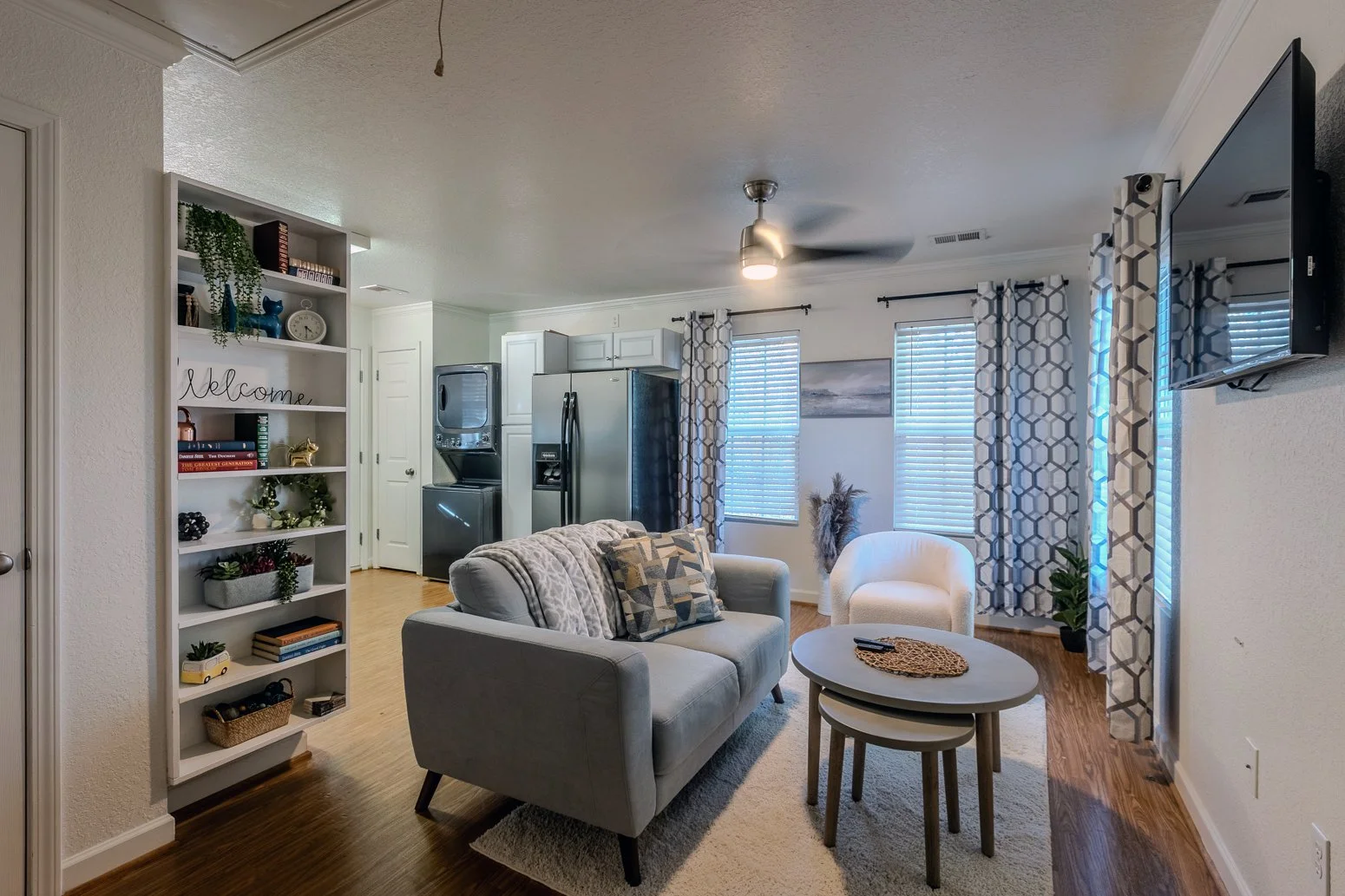 Living room with gray sofa, white armchair, wooden coffee tables, wall-mounted TV, curtains, and windows with blinds.