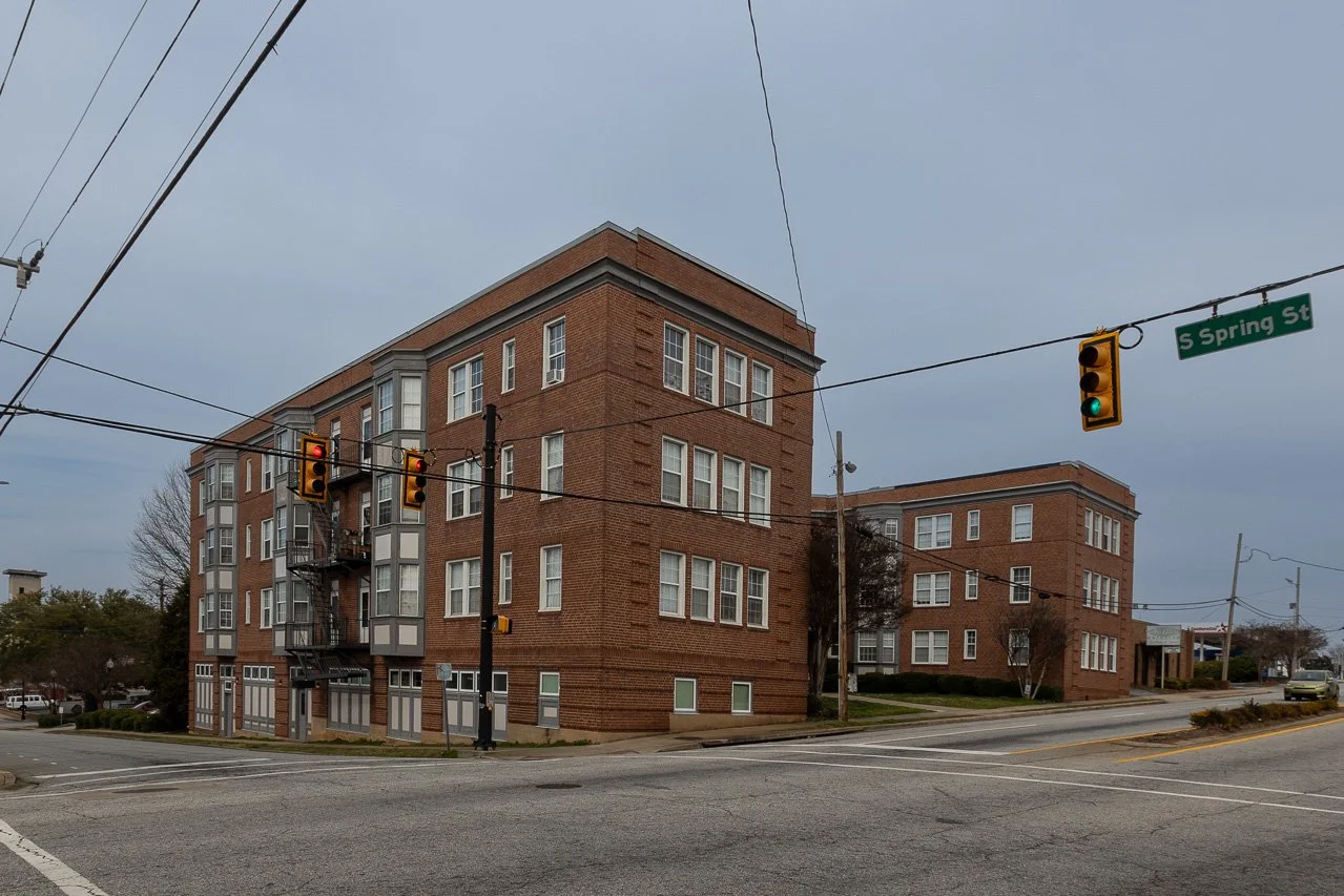 A street view showing a four-story brick apartment building at the corner of an intersection with traffic lights and the street sign 'S Spring St'. The sky is overcast.