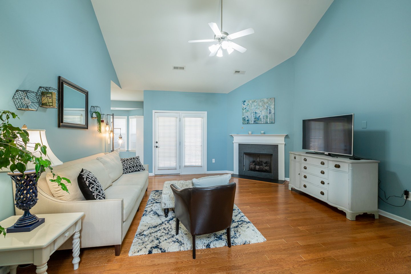 Living room with blue walls, white sofa, and a fireplace, hardwood floors, a television on a white dresser, and double doors with blinds.