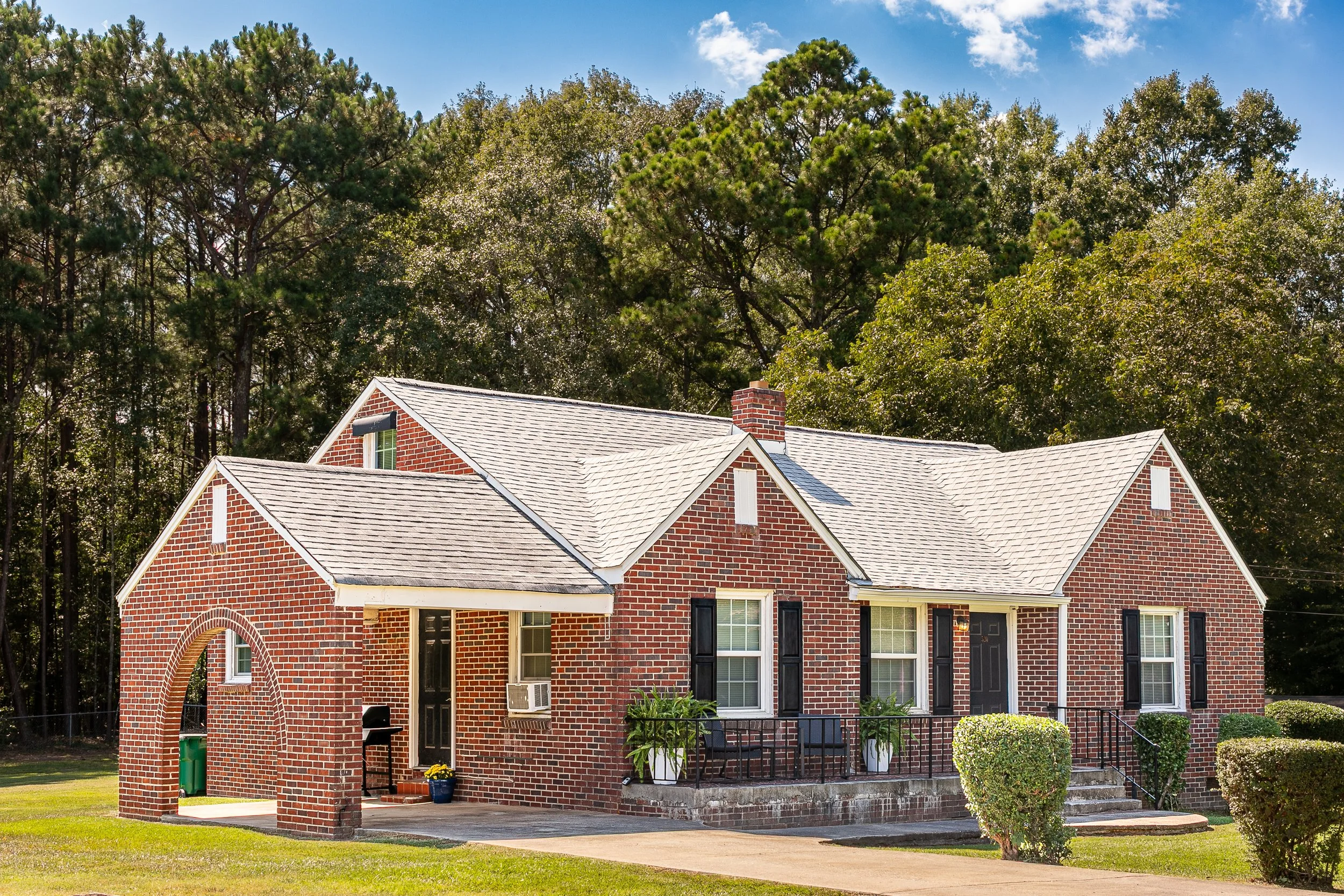 A brick house with a pitched roof, front porch with potted plants, black shutters, and a small arched brick structure on the side, surrounded by green lawn and bushes, with tall trees in the background and a partly cloudy sky.