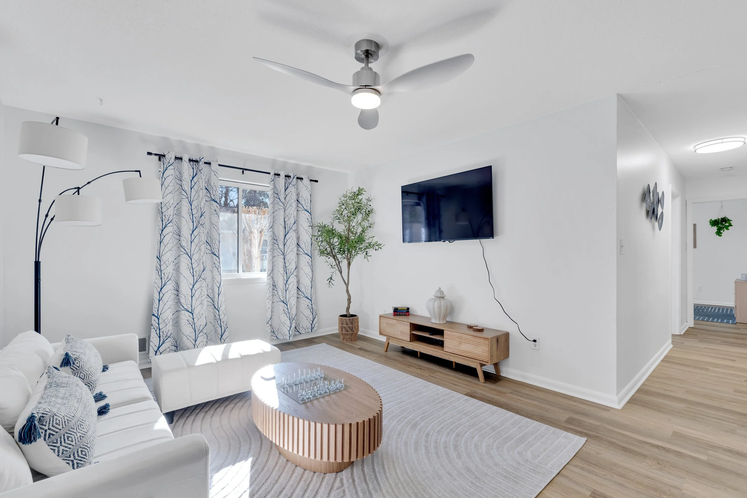 Bright living room with white walls, a large window with blue and white curtains, a white sofa with patterned pillows, a wooden coffee table with chess pieces, a tall potted plant, a wooden TV stand with a white vase, a wall-mounted flat-screen TV, a