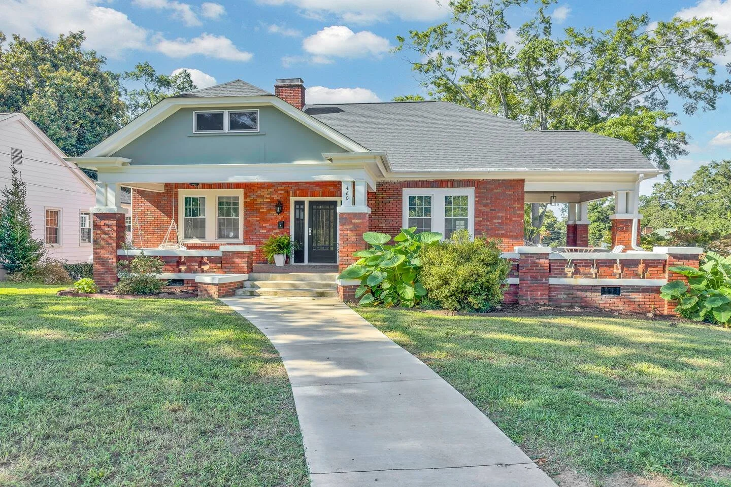 Bright red brick house with a large front porch, two chairs, lush greenery, a curved sidewalk leading to the front steps, and a well-maintained lawn, under a partly cloudy sky.