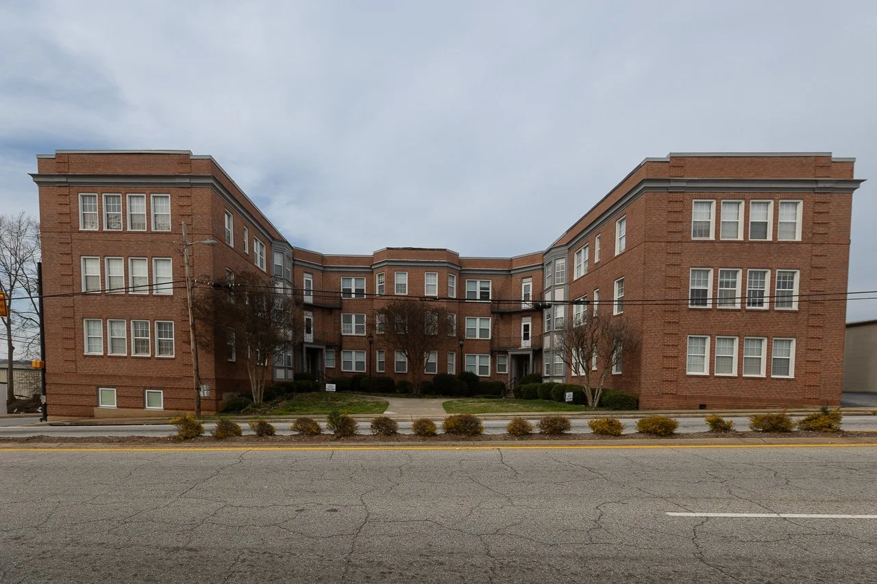 A three-story brick apartment building with multiple windows and small trees in front, sitting behind a street with a double yellow line and small bushes along the sidewalk.
