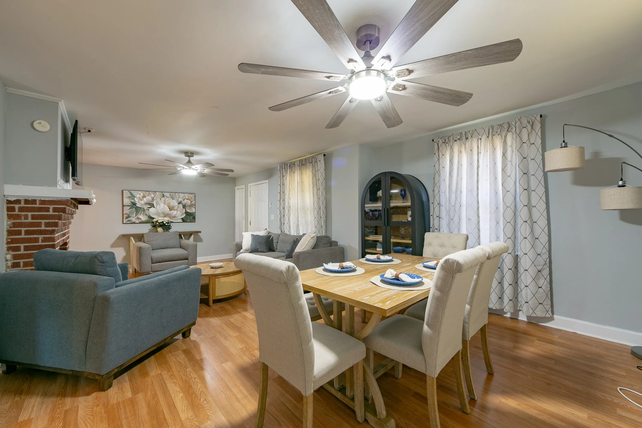 A spacious living and dining area with hardwood floors, light blue walls, and large windows with patterned curtains. The ceiling has two ceiling fans. The dining table is set with six plates and napkins, and beige upholstered chairs surround it. The 