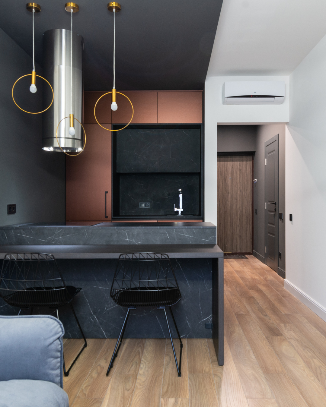 Modern kitchen with black marble countertop, black wire chairs, pendant lights, cabinetry with wood and black finishes, and a wooden door.
