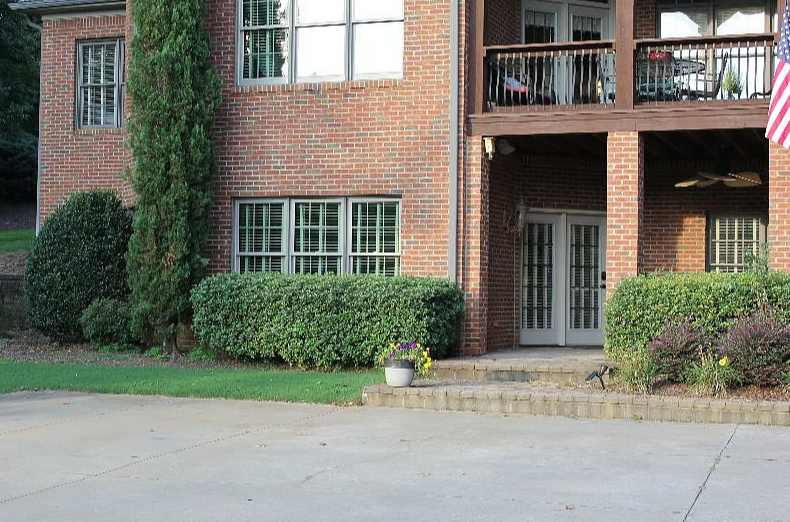 Exterior view of a brick apartment building with stairs leading to glass door entrance, surrounded by green bushes and potted plants, with an American flag on the balcony.
