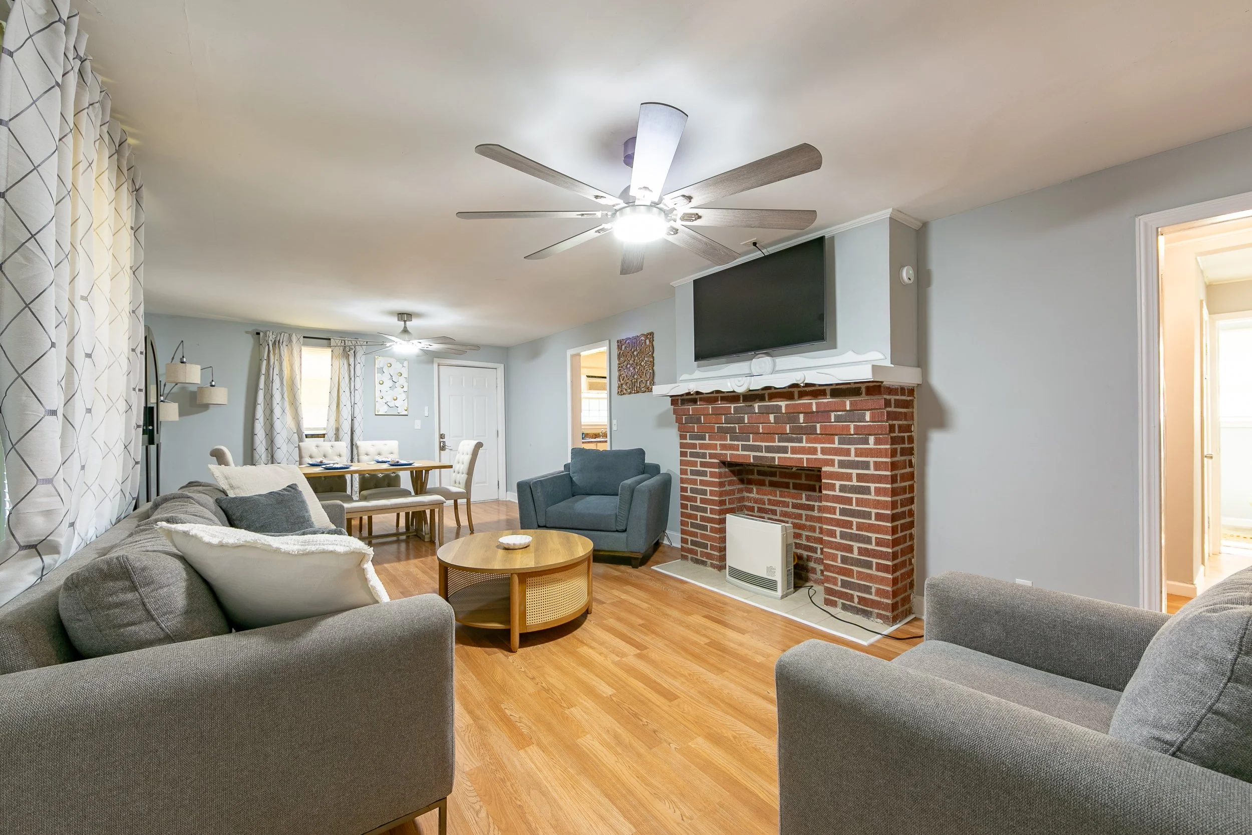 Living room with gray and beige furniture, a brick fireplace with a TV above, hardwood floors, and light-colored walls.