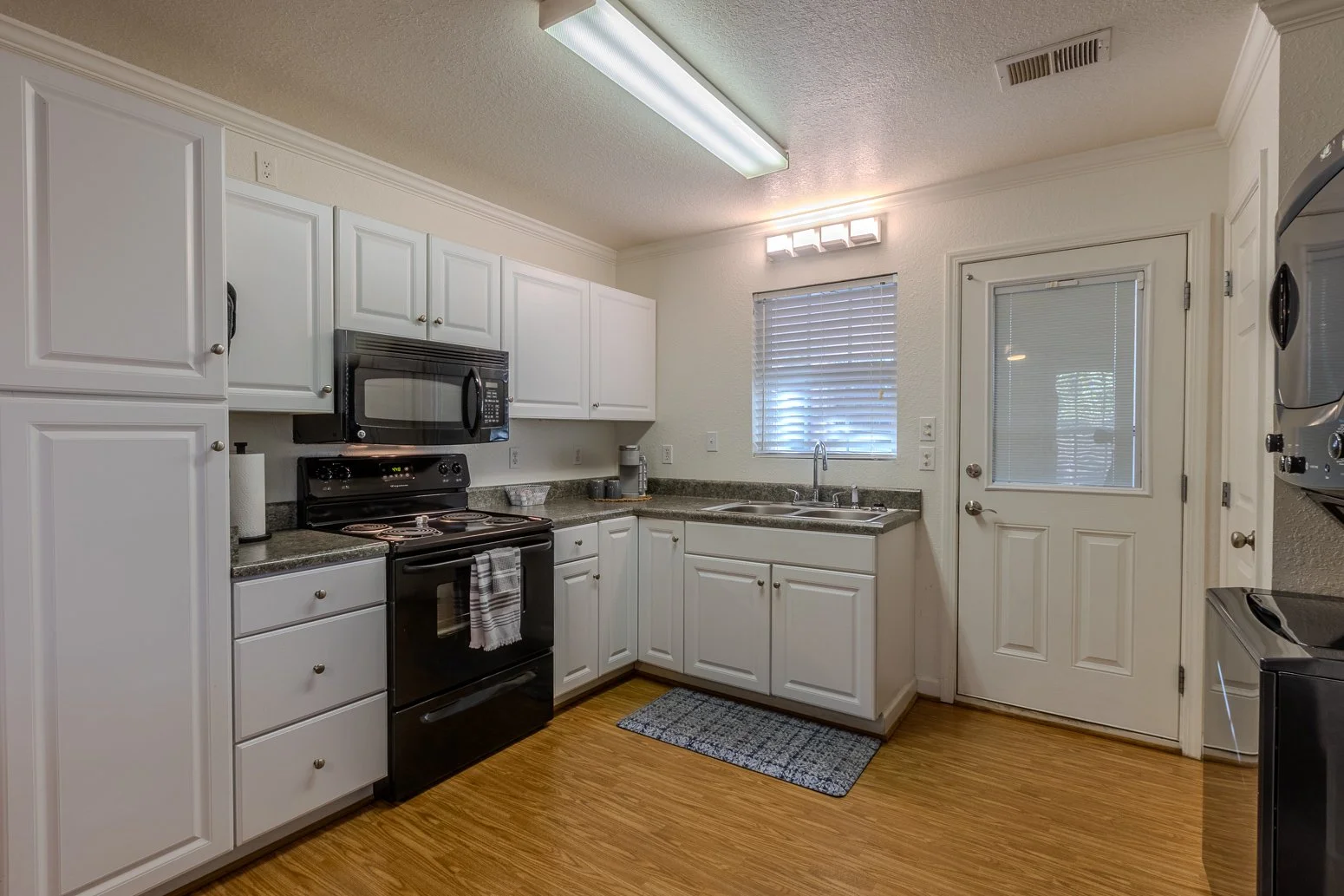 Kitchen with white cabinets, a black stove and microwave, granite countertops, a sink under a window, wooden flooring, and a door with a window leading outside.