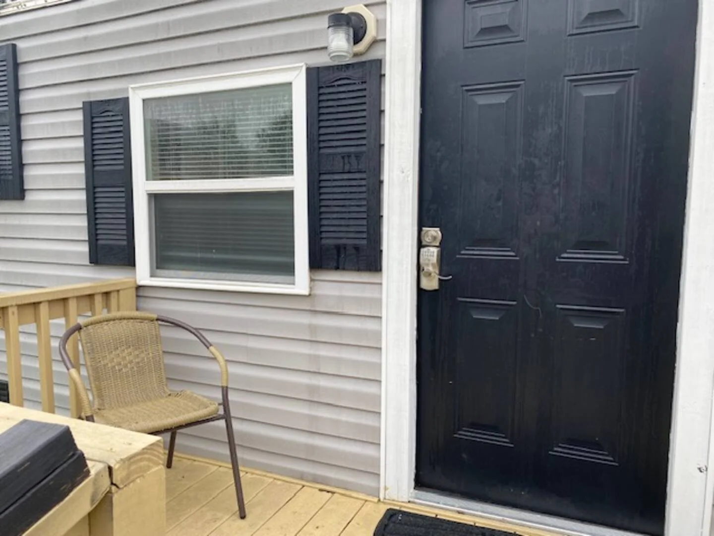 A wooden porch with a beige wicker chair next to a cardboard box, in front of a house with gray vinyl siding, a window with black shutters, and a black door.