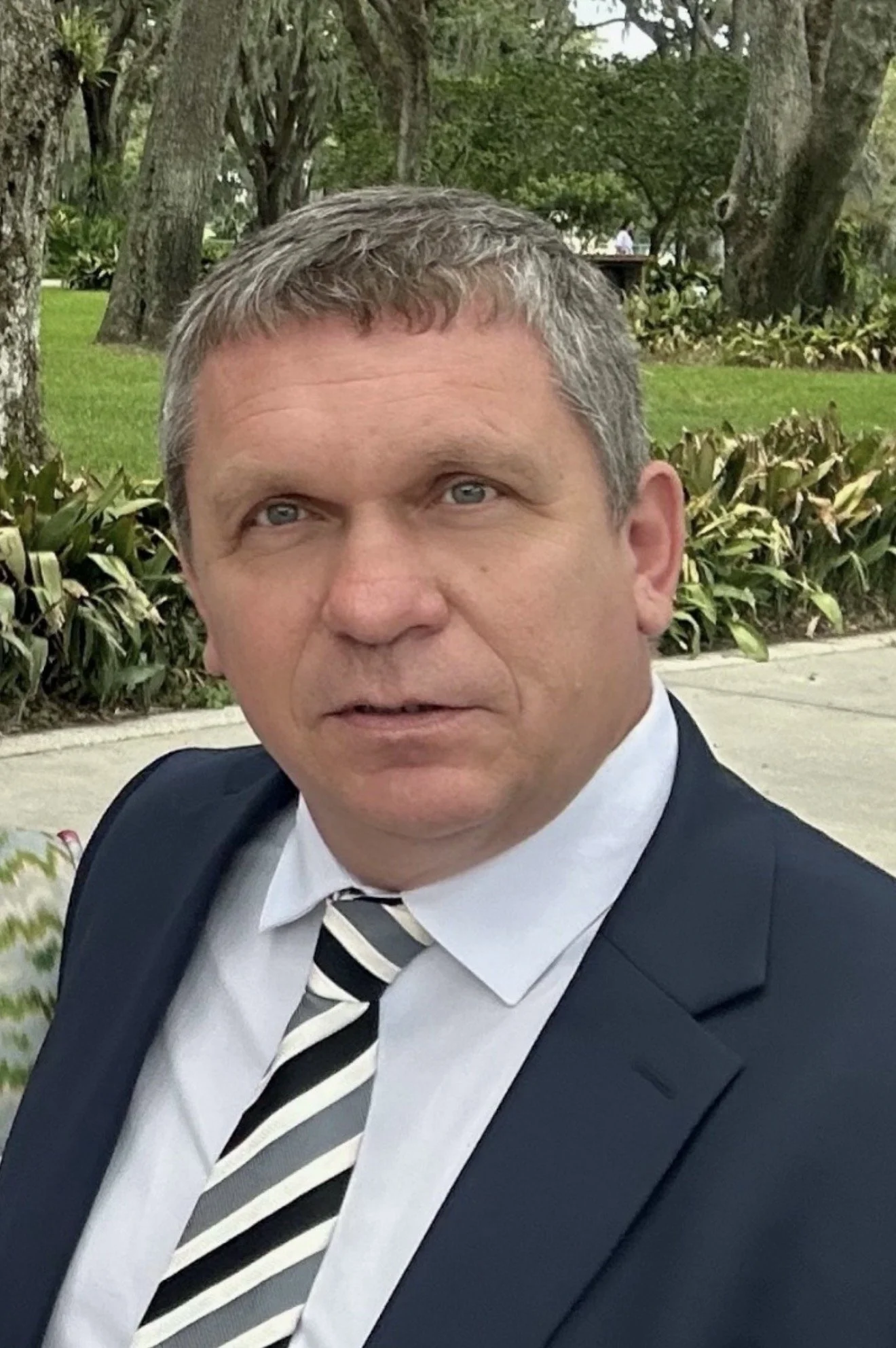 A man in a suit and striped tie outdoors, with trees and green grass in the background.