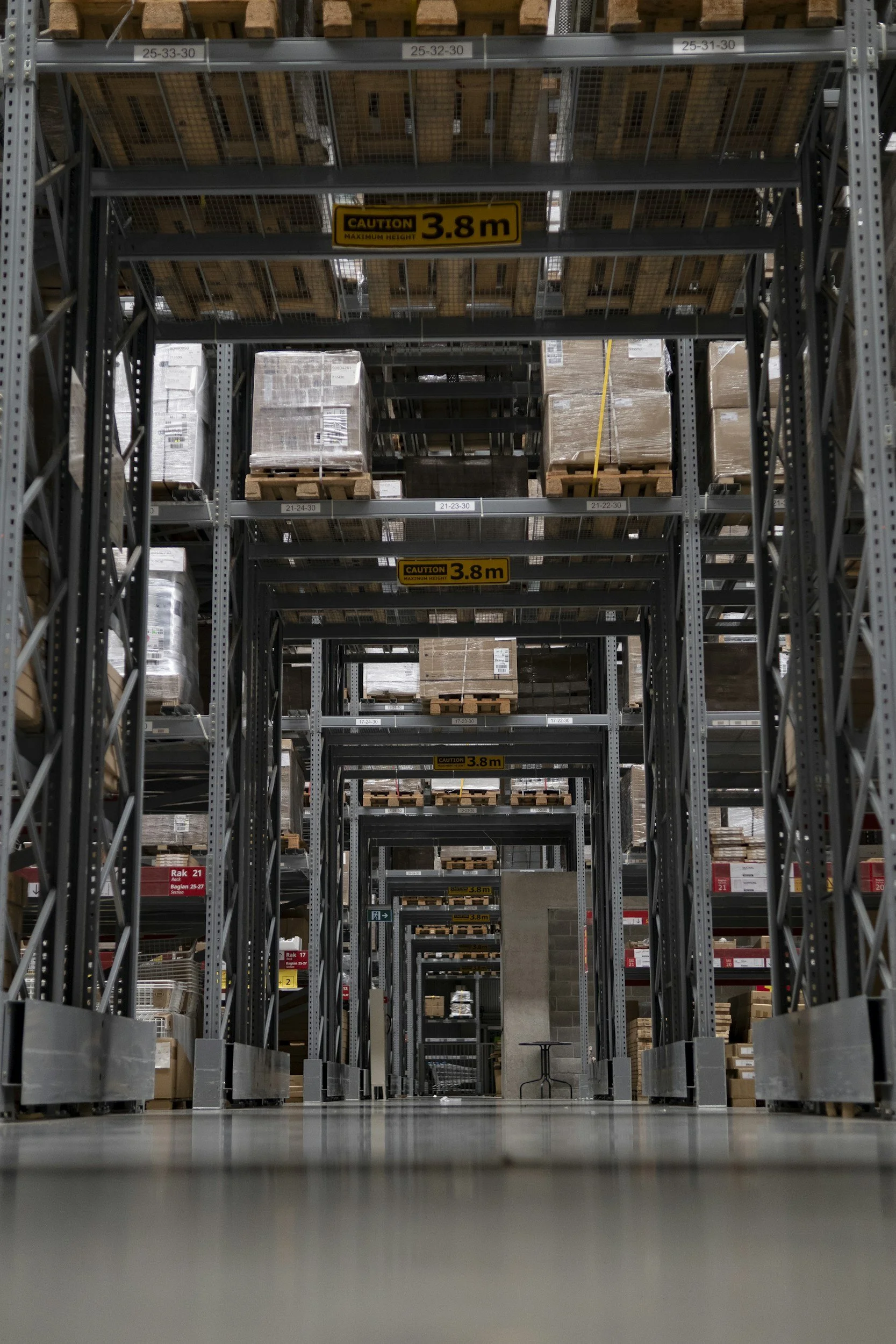 View of warehouse storage shelves filled with stacked boxes and pallets, with yellow signs indicating a maximum height of 3.8 meters.