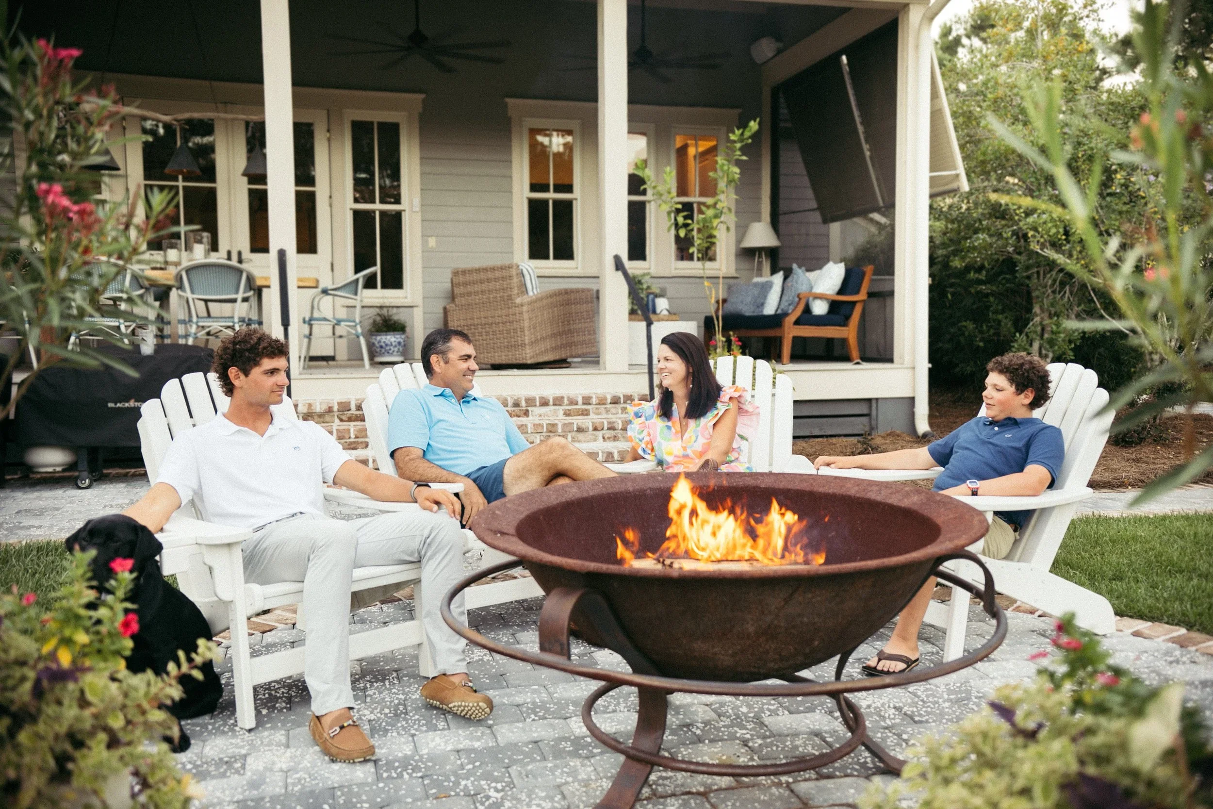 Family of four sitting around a fire pit in a backyard, with a woman and man laughing and children relaxing in Adirondack chairs.