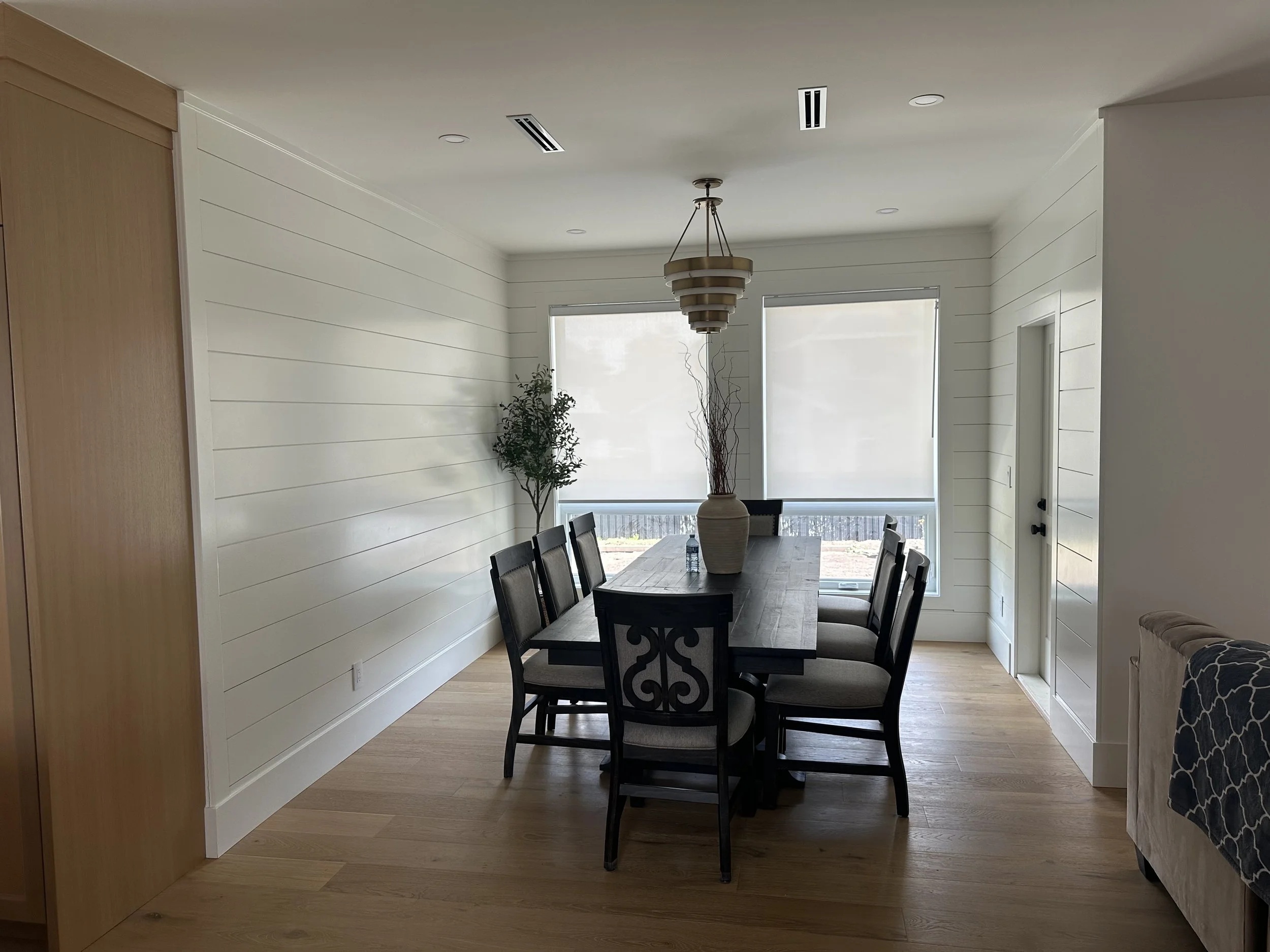 Dining room with wooden table, eight chairs, light wood flooring, white shiplap walls, large windows with blinds, potted plant, and decorative vase with twigs in the center of the table.
