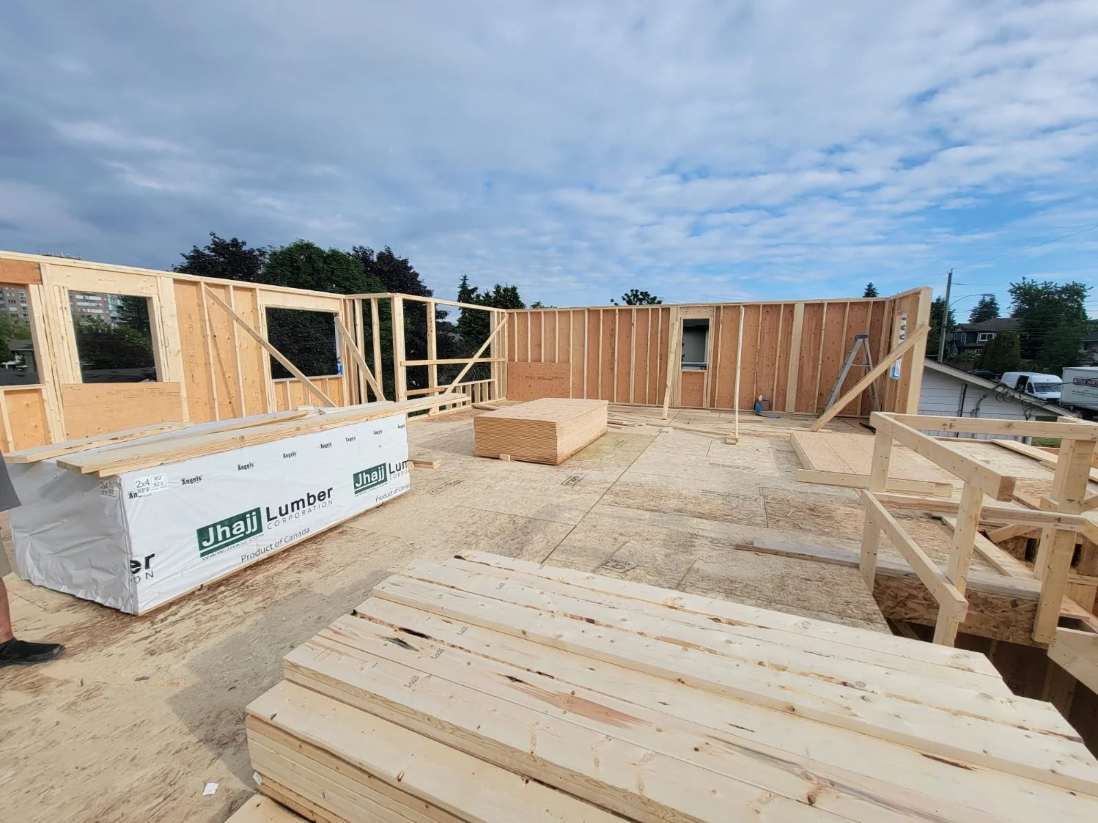 Construction site with wooden framing inside a building under construction, stacks of lumber, and a partially built wall. Sky with clouds visible in the background.
