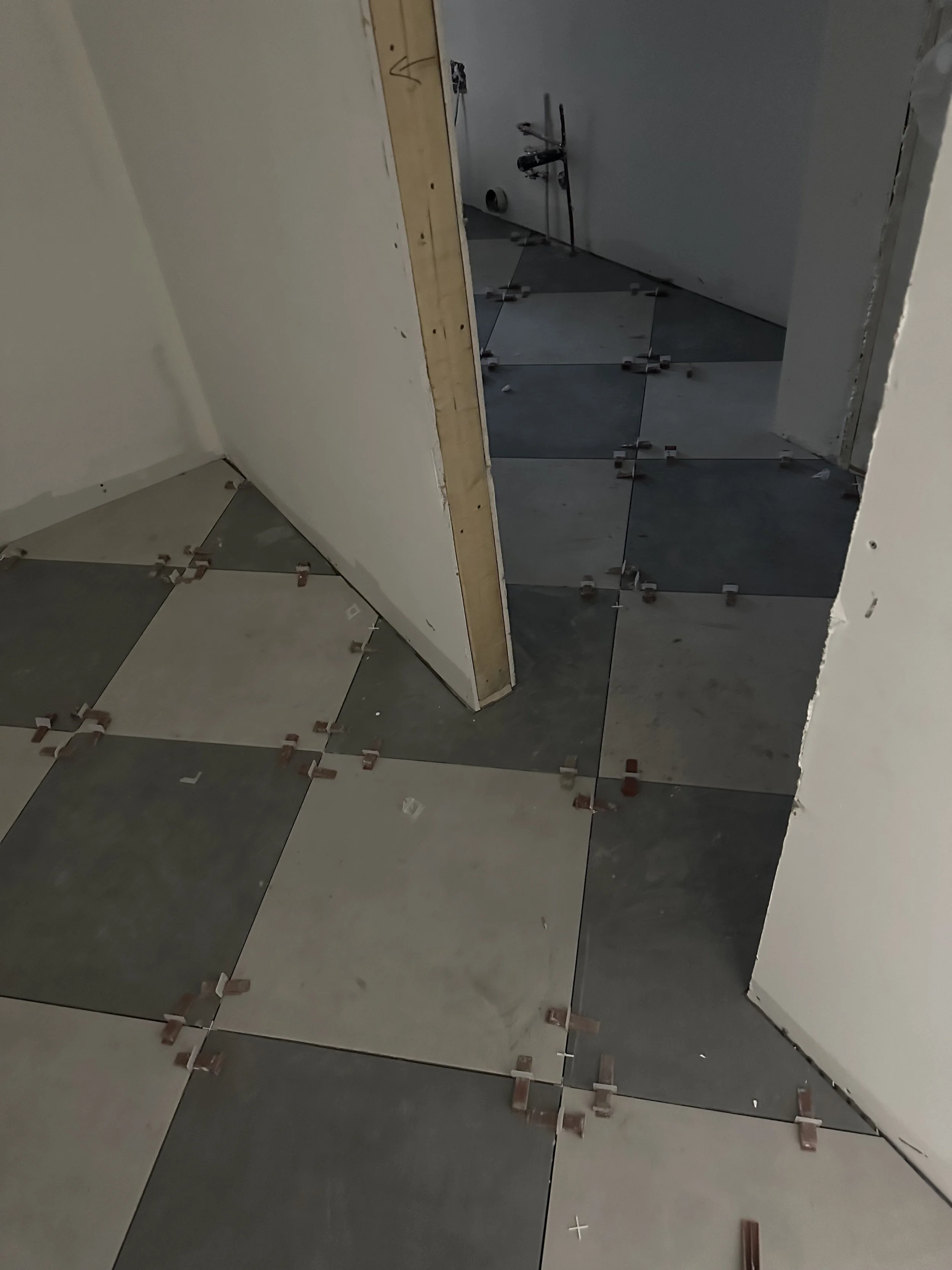 Interior of a room under construction with a partially finished tiled floor featuring gray and beige tiles, some tiles are cut and bricks are used as spacers between tiles.