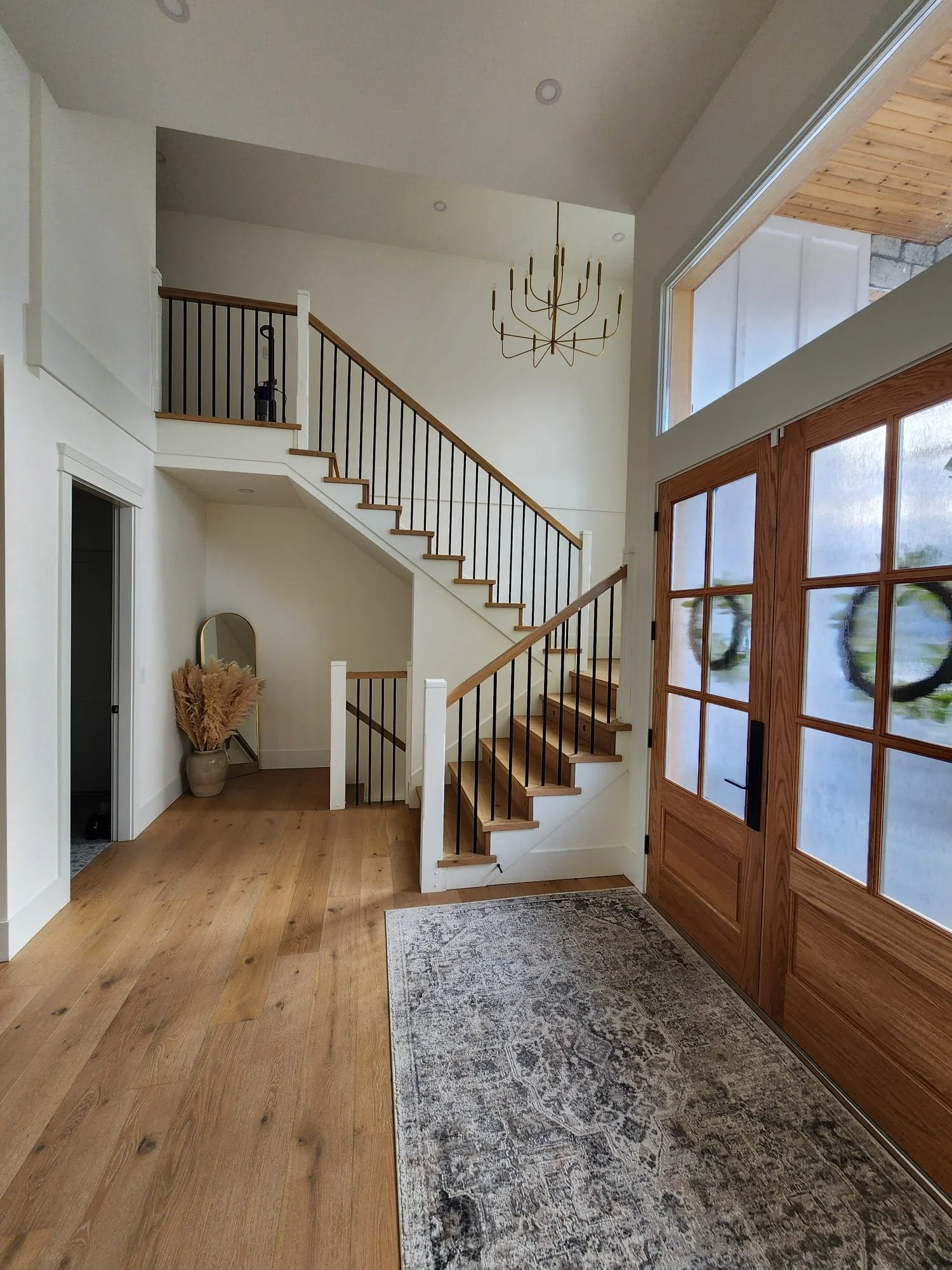 The image shows a modern home entryway with a large wooden front door with glass panels, a light-colored hardwood floor, and a staircase with wooden steps and black metal railing leading upstairs. There is a hanging gold chandelier, a small mirror ne