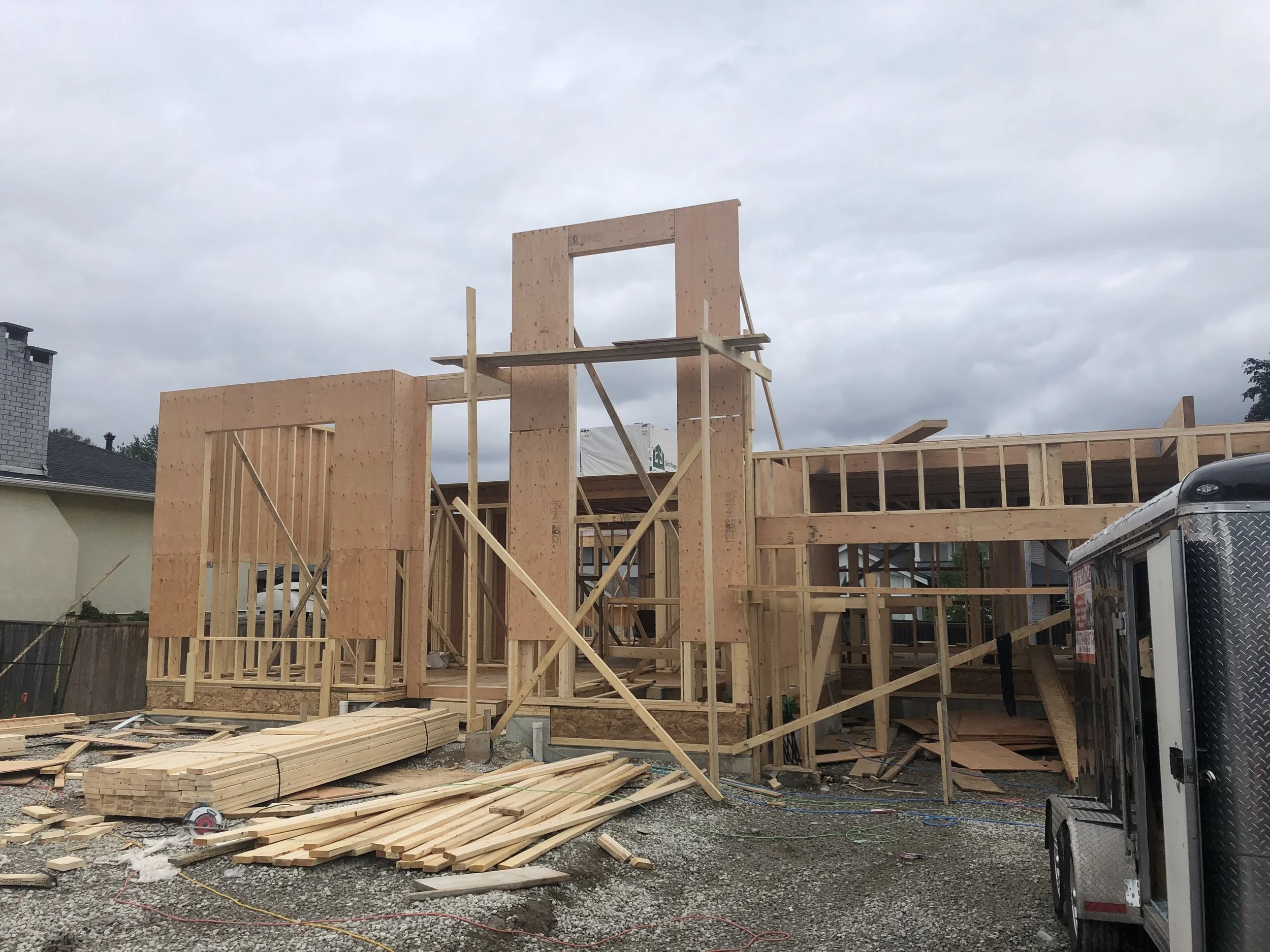 Construction site with wooden framing for a house, showing partially built walls and scaffolding, and construction materials on the ground.