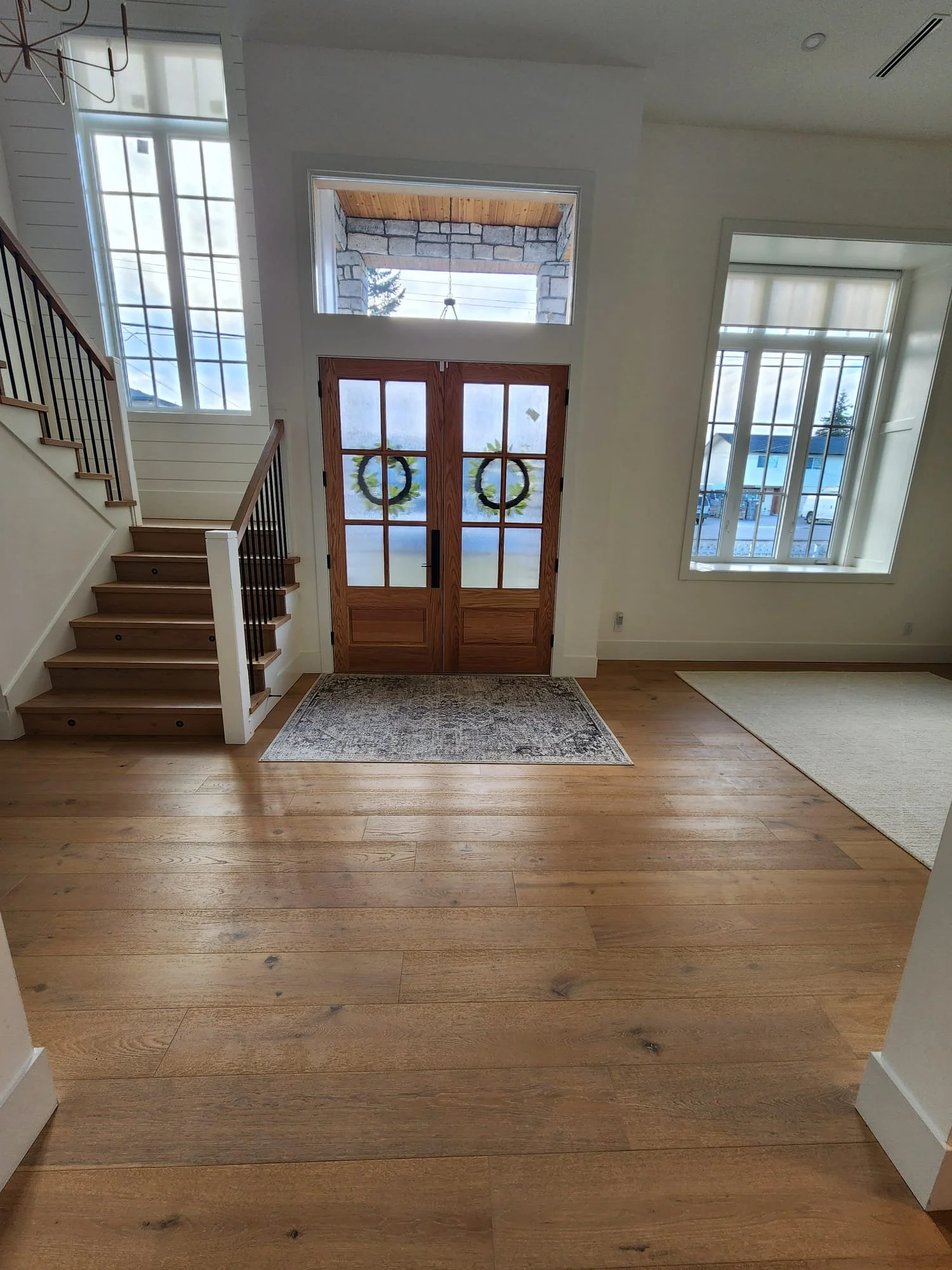 Entryway of a house with wooden front doors decorated with wreaths, large windows on either side, a staircase with wooden steps and black railing on the left, hardwood flooring, and two area rugs, one in front of the door and one on the right side.