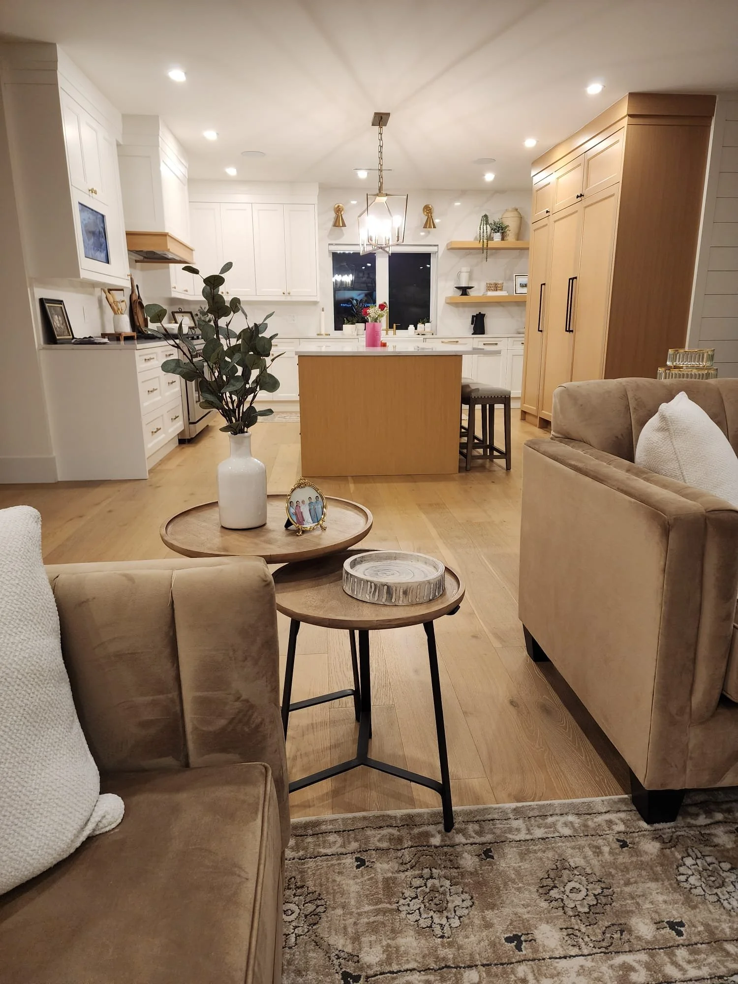 Open-concept living room and kitchen with beige sofas, wooden tables, and modern decor, illuminated by ceiling lights and a central hanging light fixture.