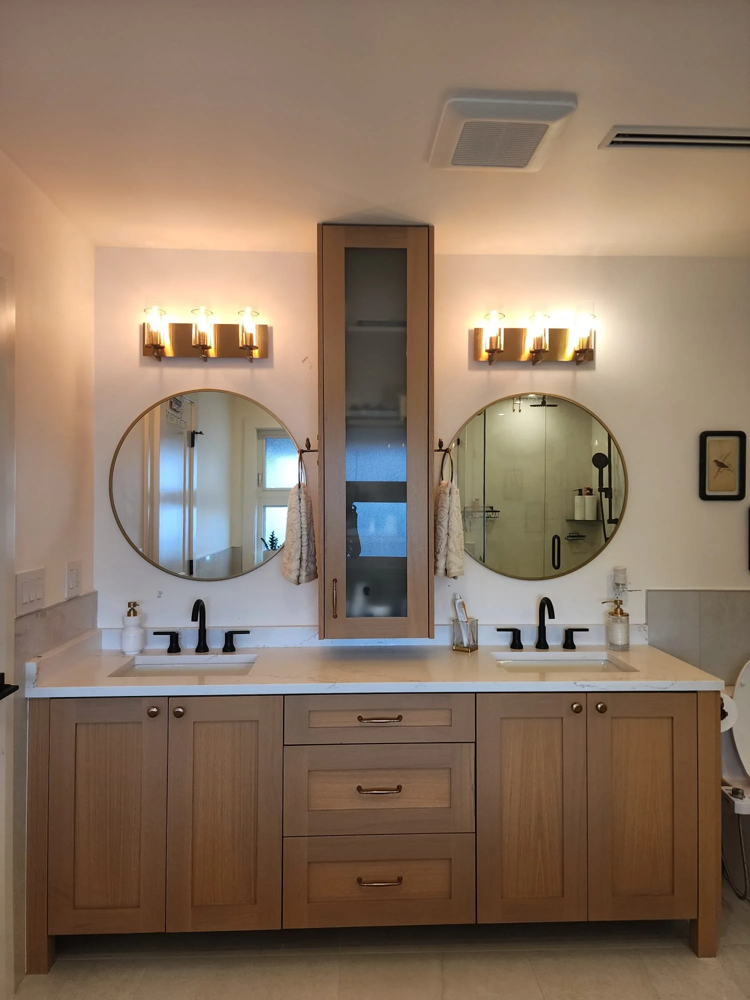 Double bathroom vanity with two round mirrors, black faucets, and wooden cabinetry. There are light fixtures above each mirror, and a central cabinet between the mirrors. Towels hang on hooks beside the mirrors, and soap dispensers are on the counter
