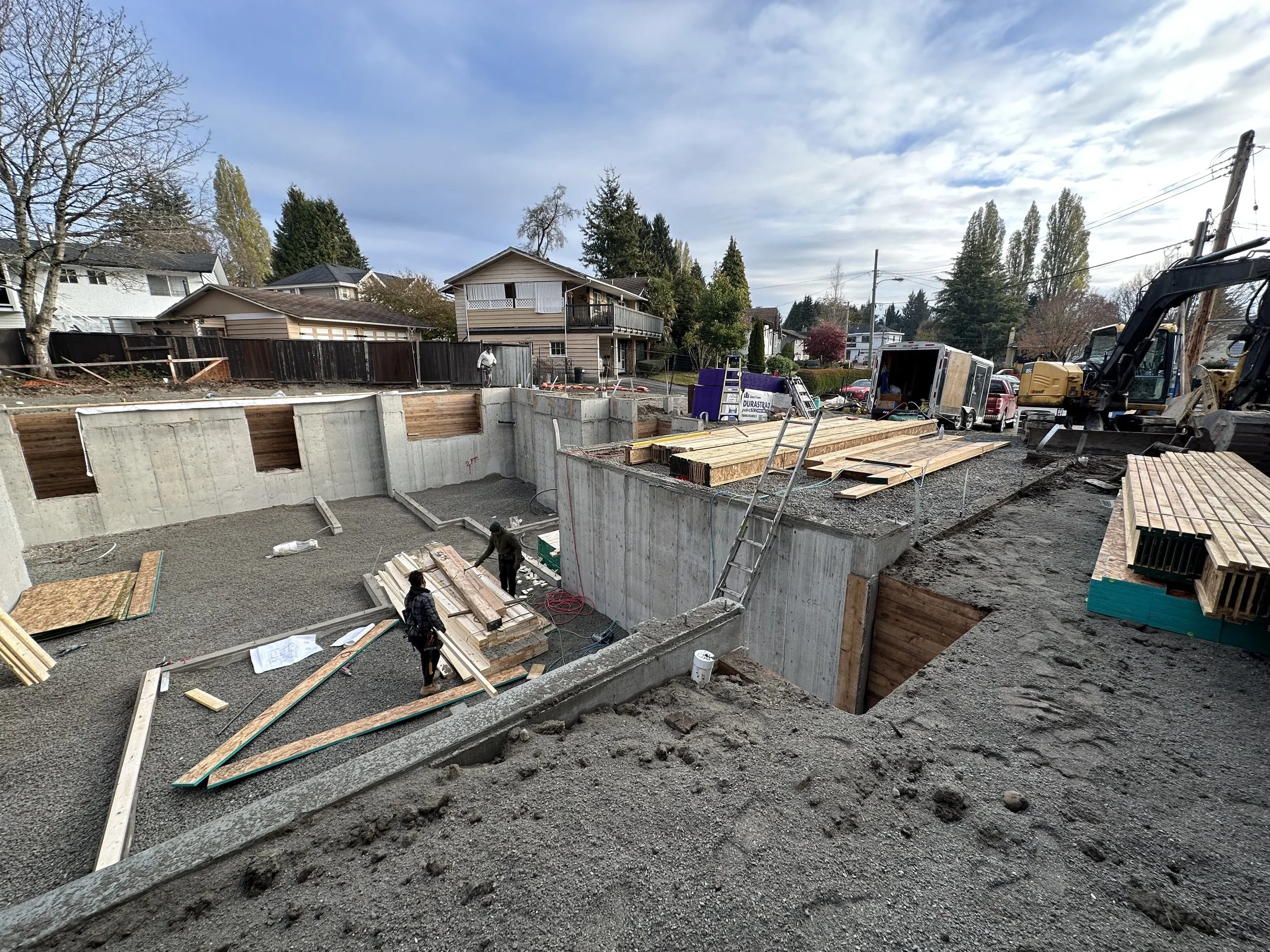 Construction site with a partially built concrete pool, construction workers, wooden planks, construction vehicles, and equipment, residential houses in the background, and a cloudy sky.