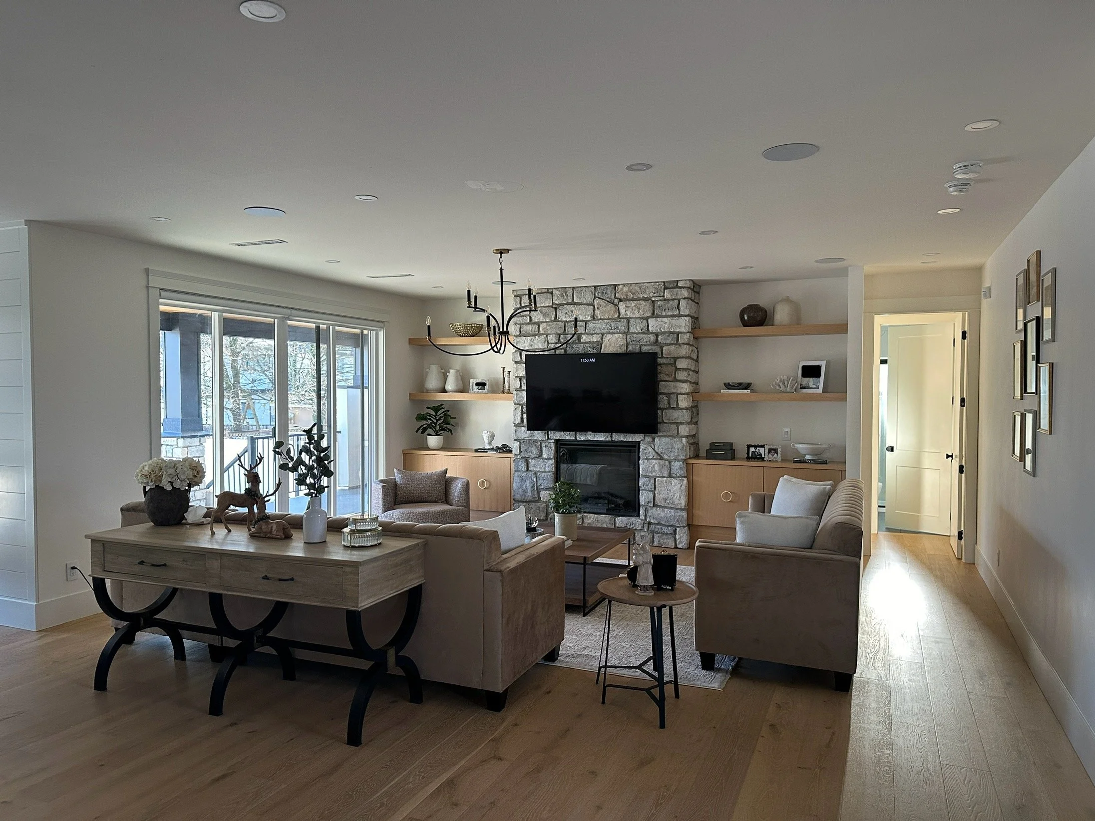 Living room with large sliding glass door, stone fireplace, and contemporary furniture including chairs, a sofa, and a console table with decorative items. Shelves with decor and a ceiling chandelier visible.