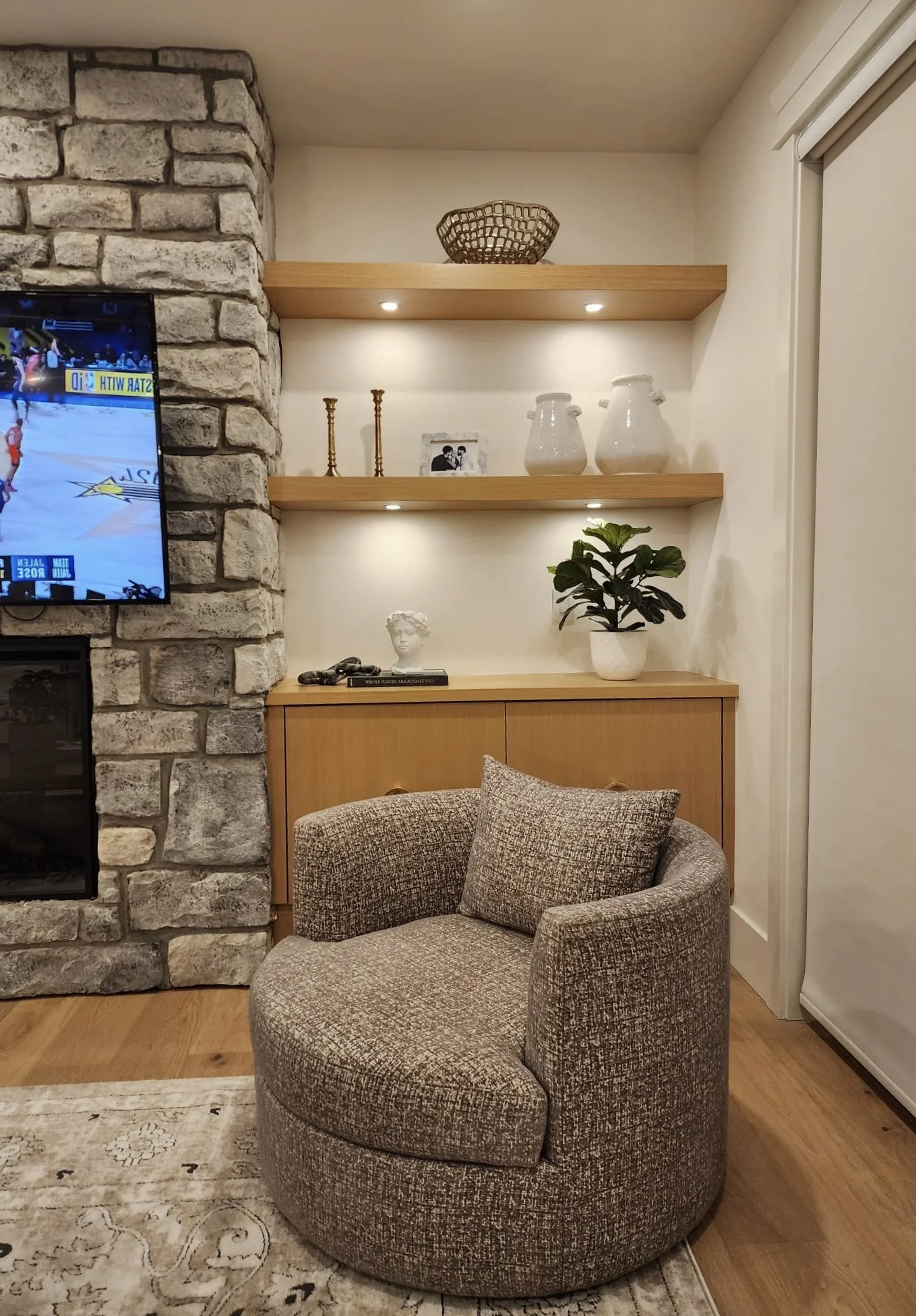 Cozy living room corner with a brown upholstered armchair, a wooden sideboard decorated with books, a sculpture, a plant in a white pot, and decorative items on wall-mounted wooden shelves with recessed lighting, stone fireplace on the left, and a TV