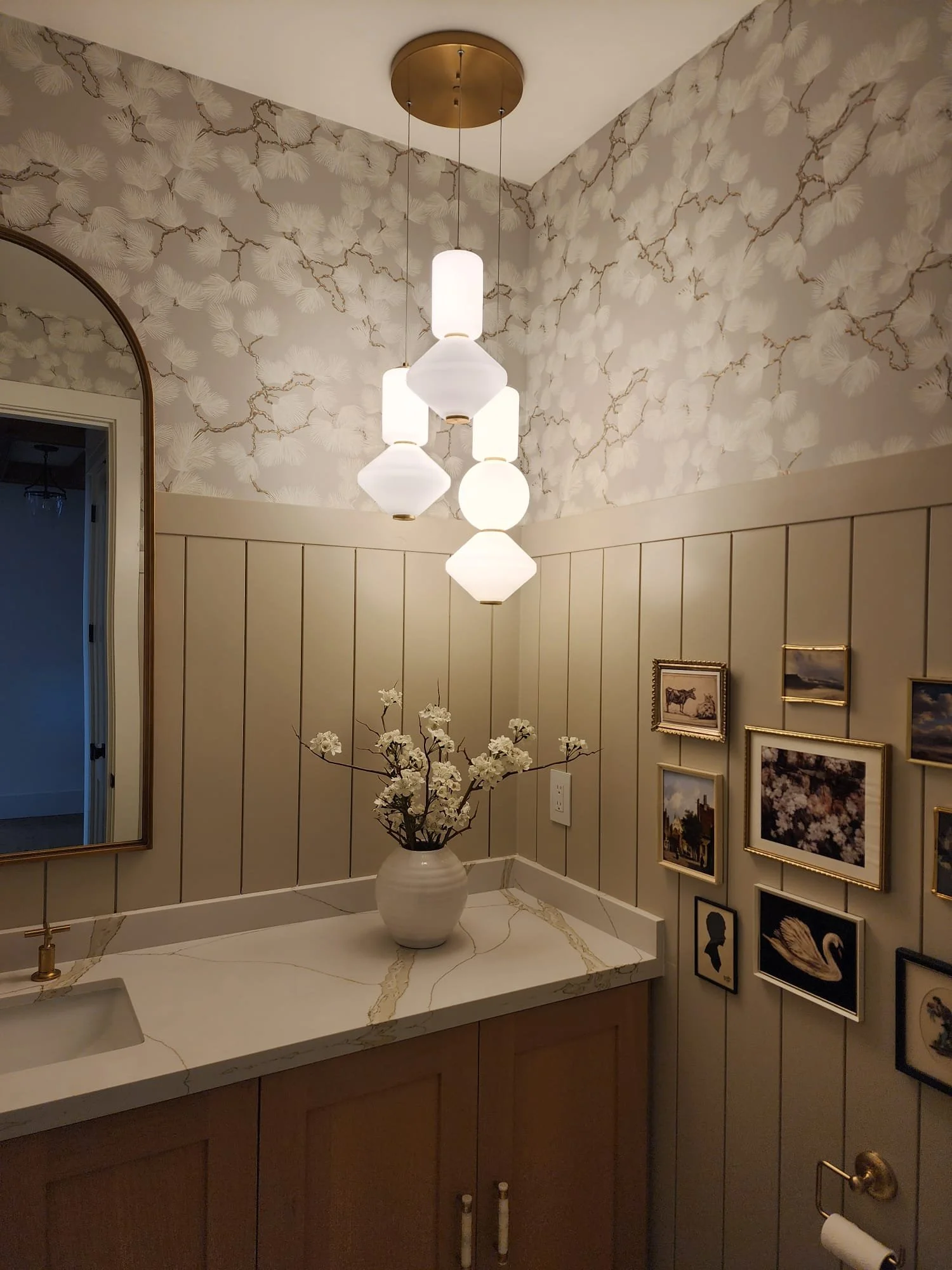 A bathroom with beige paneled walls, a white marble countertop with gold veins, a white vase with white flowers, a golden towel holder, a large framed mirror, and a gallery wall of framed pictures. There is a modern chandelier hanging from the ceilin